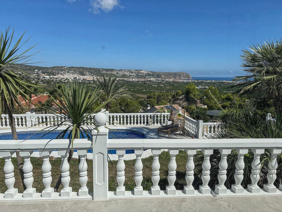 Terraza elevada con balaustrada blanca que ofrece vistas panorámicas al valle y al mar Mediterráneo, con la zona de piscina y palmeras en primer plano.