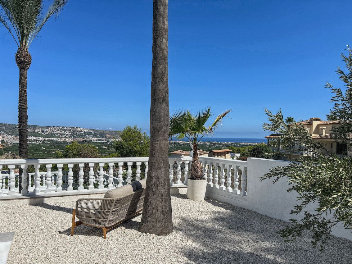 Terraza exterior con suelo de grava blanca, mobiliario de diseño y vistas panorámicas al mar y al valle, enmarcada por palmeras y una balaustrada clásica.