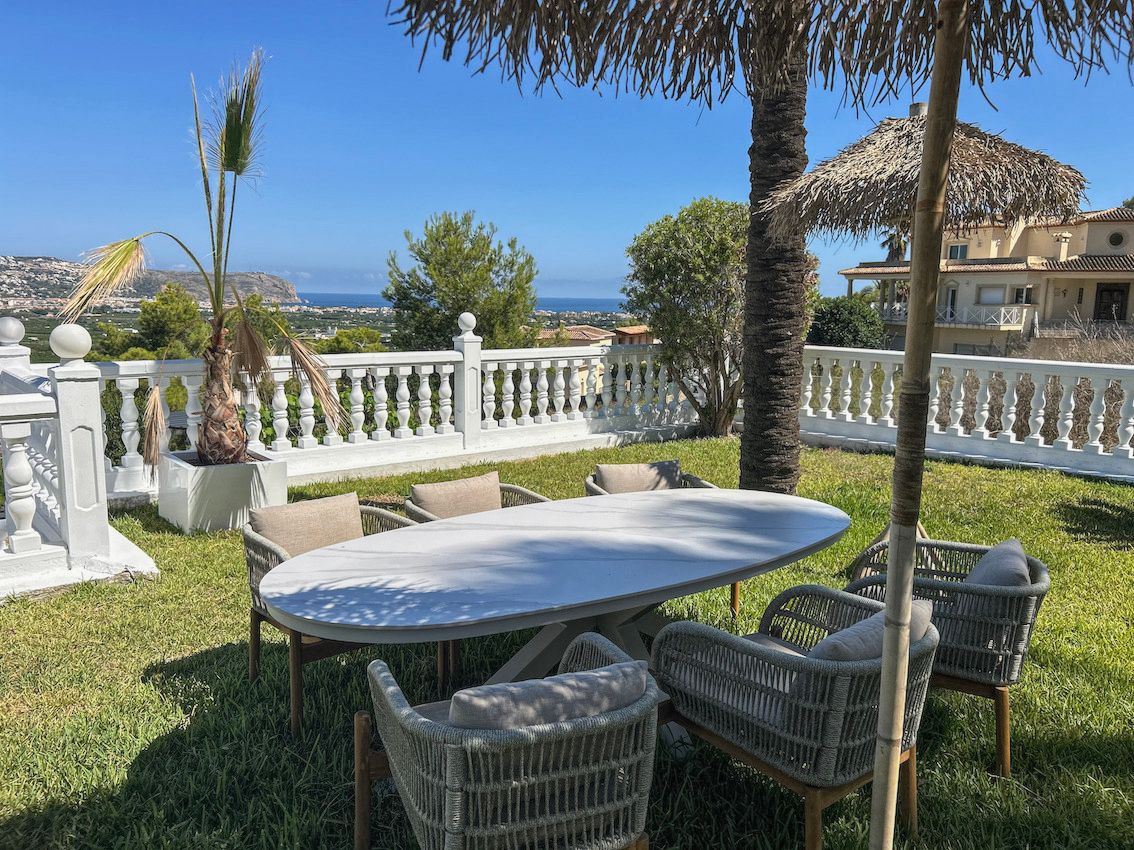 Zona de comedor exterior sobre césped con mesa ovalada y sillas de diseño, rodeada de una balaustrada blanca con vistas panorámicas al mar y al valle.