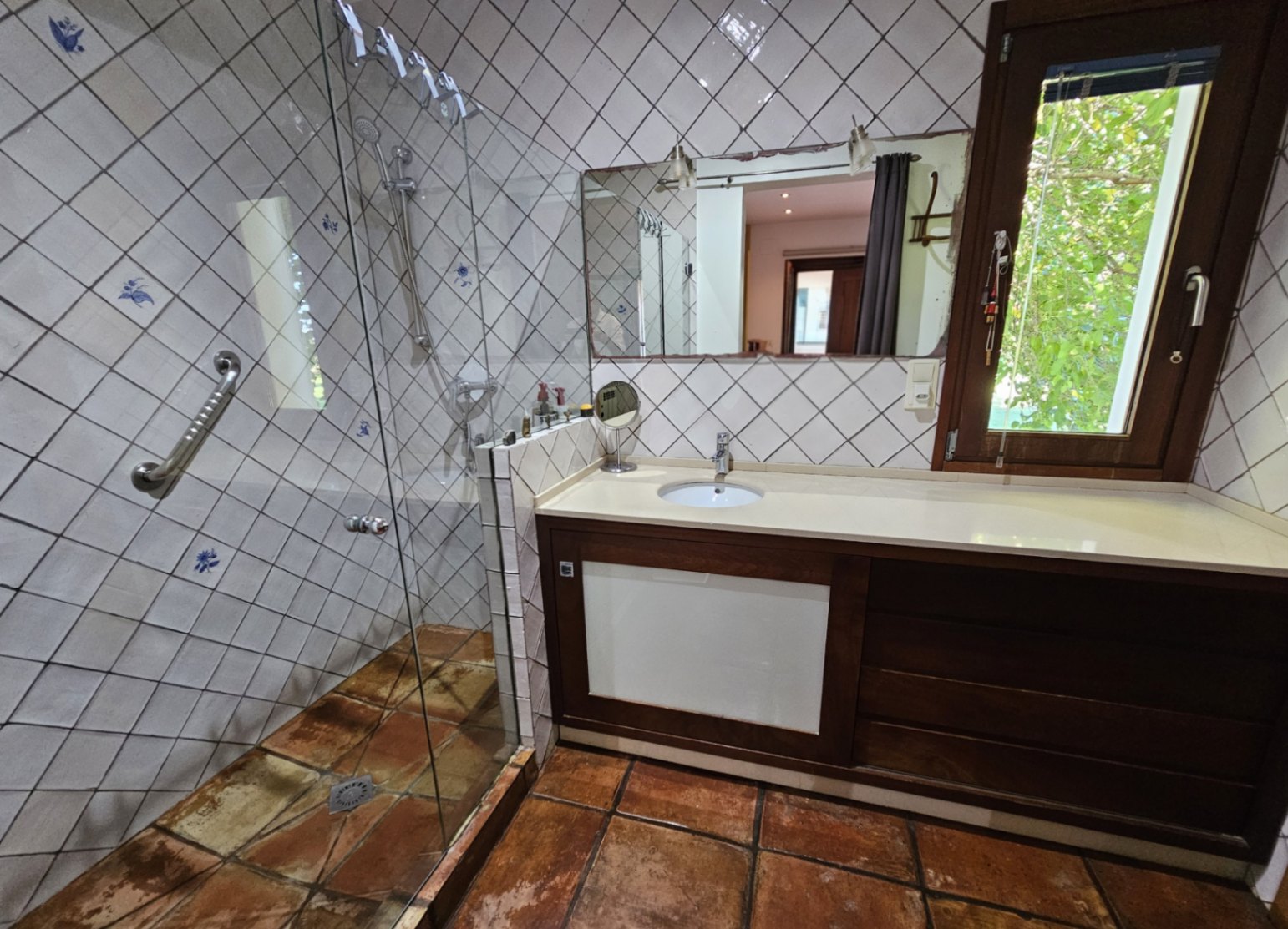 Baño con azulejos pintados a mano, ducha de obra con mampara de cristal y suelos de barro artesanal. Incluye un amplio mueble de madera y ventana con vistas al jardín.