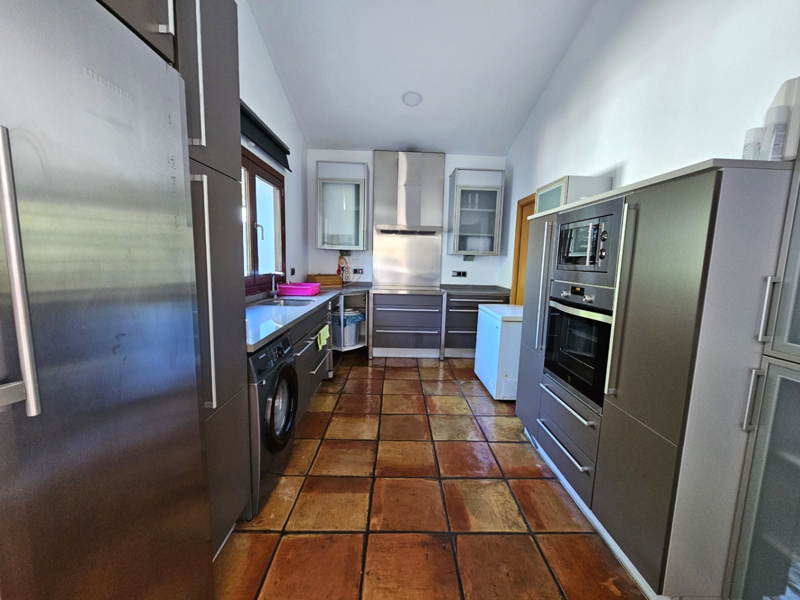 Cocina moderna con armarios en gris mate, electrodomésticos de acero inoxidable y suelos de barro hechos a mano que aportan calidez al diseño funcional.