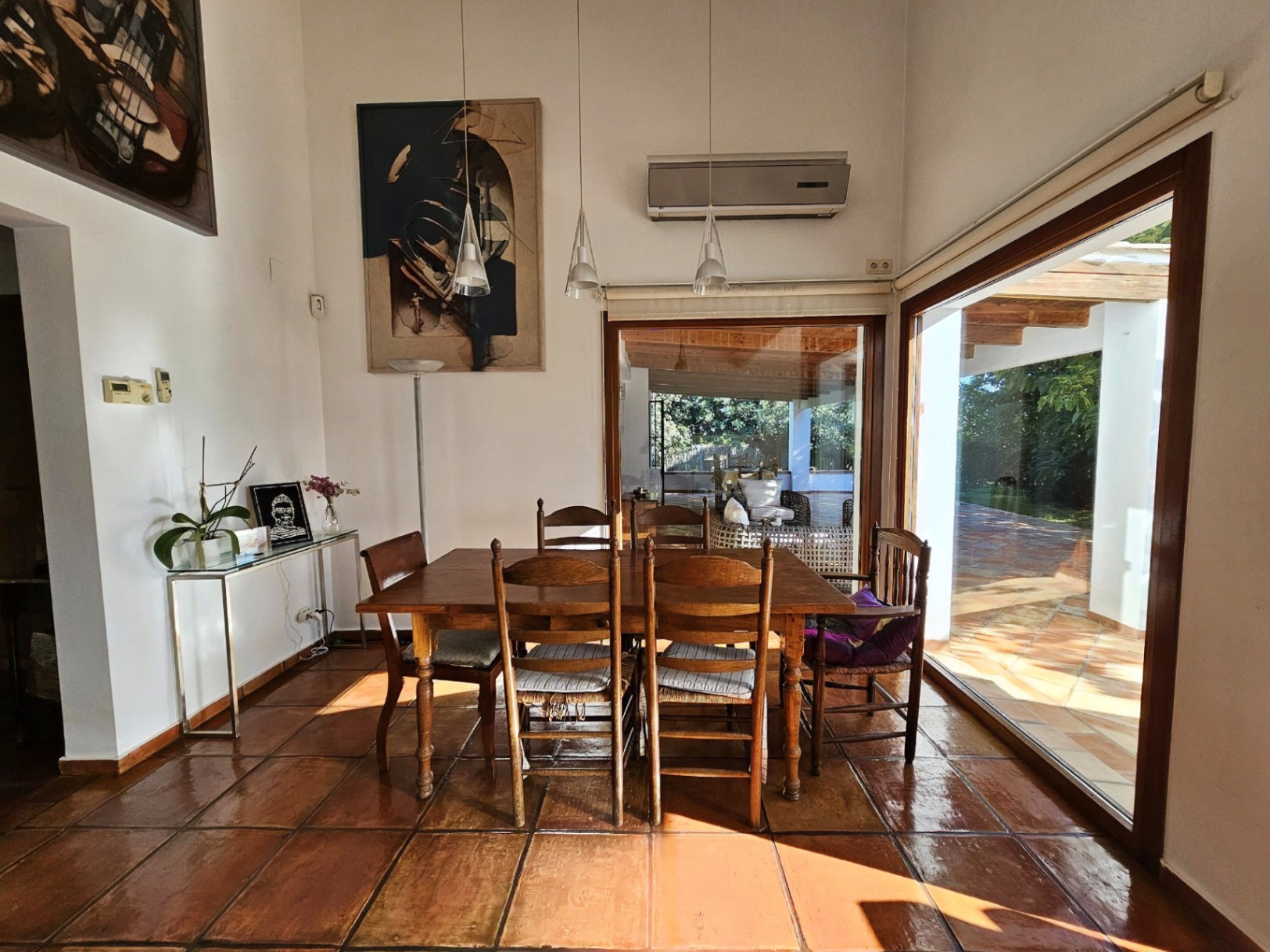 Comedor luminoso con suelos de barro hechos a mano, mesa de madera maciza y grandes ventanales que conectan visualmente con la naya y el jardín exterior.