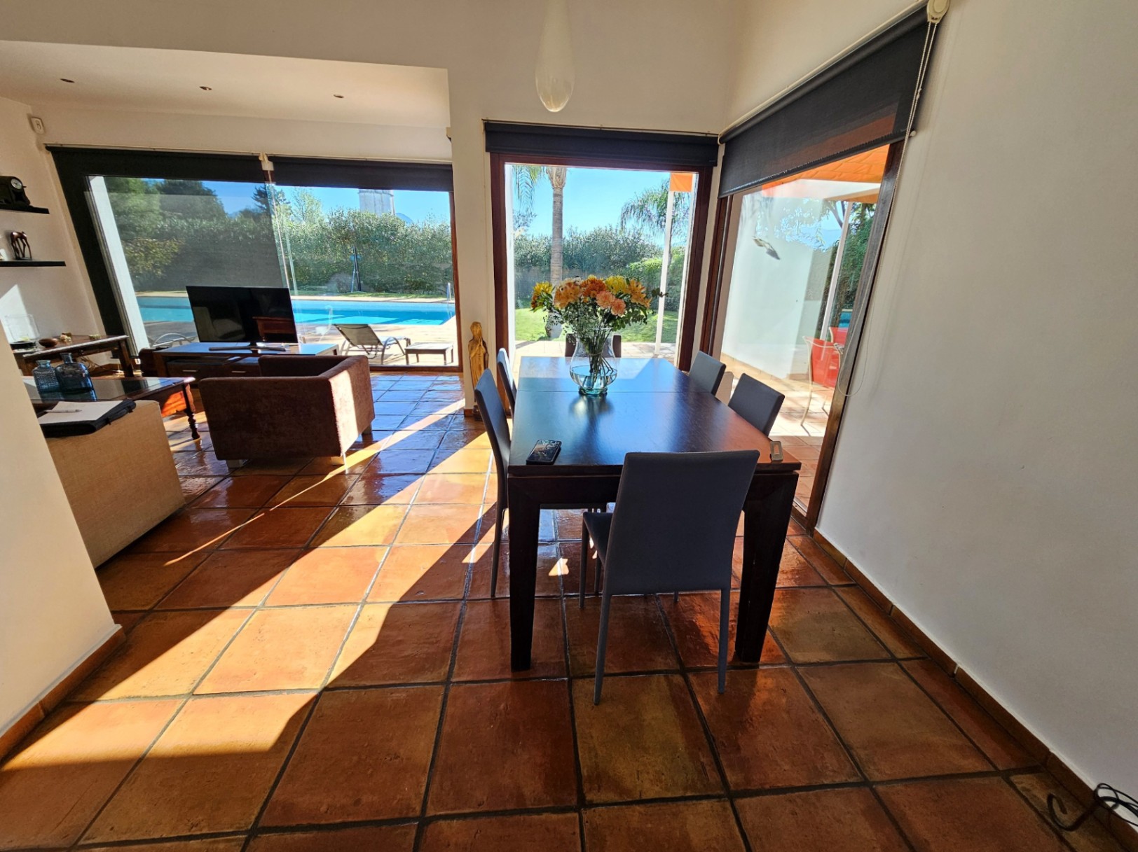 Comedor luminoso con suelos de barro hechos a mano, mesa de madera oscura y grandes ventanales que ofrecen vistas directas a la piscina y el jardín.