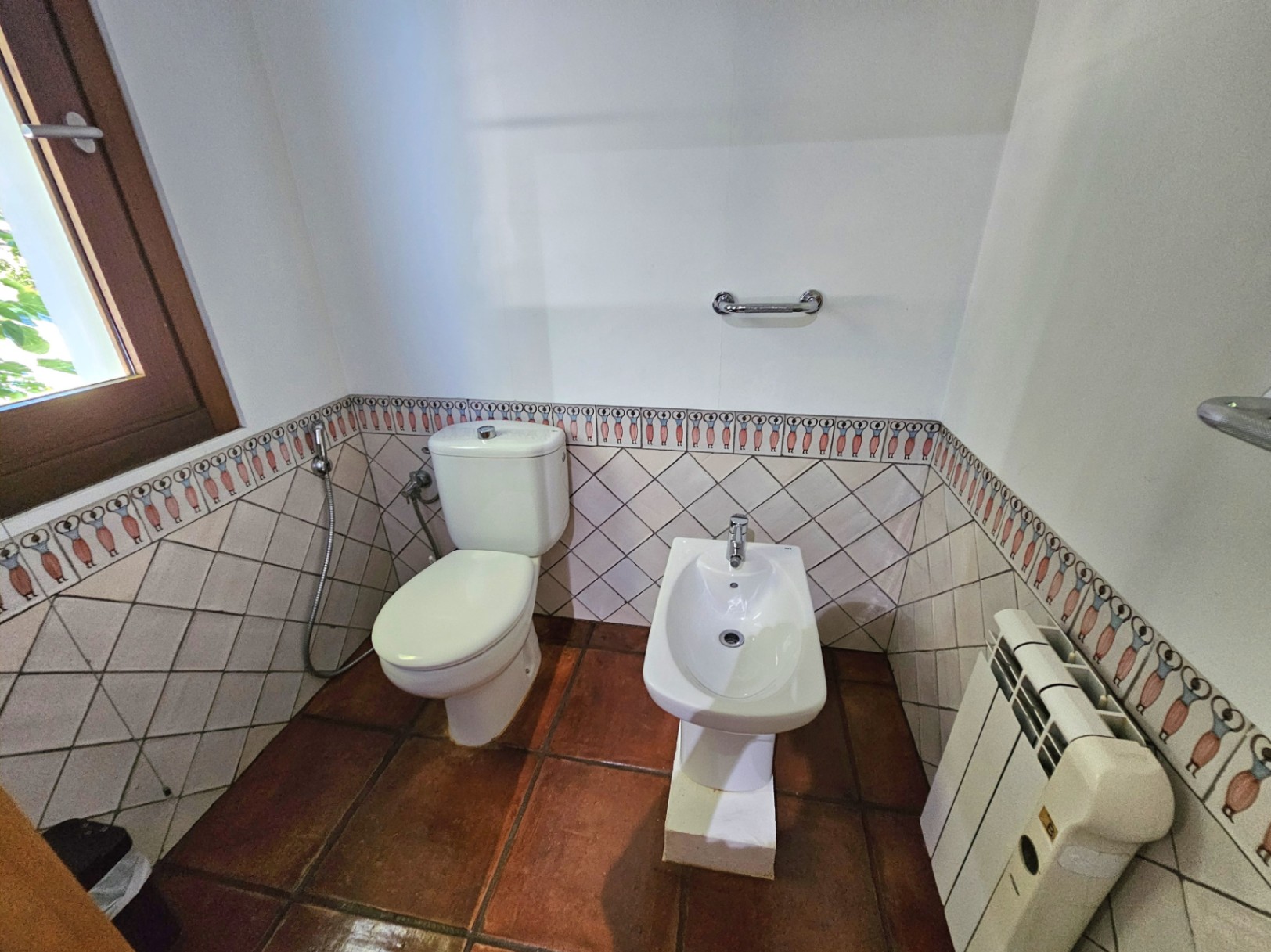 Cuarto de baño con inodoro y bidé, destacando el suelo de barro artesanal y el alicatado con cenefa decorativa pintada a mano.