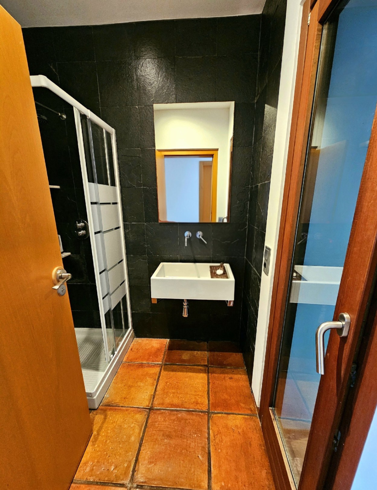 Cuarto de baño con suelos de barro hechos a mano, paredes de piedra oscura texturizada, lavabo suspendido moderno y cabina de ducha acristalada.