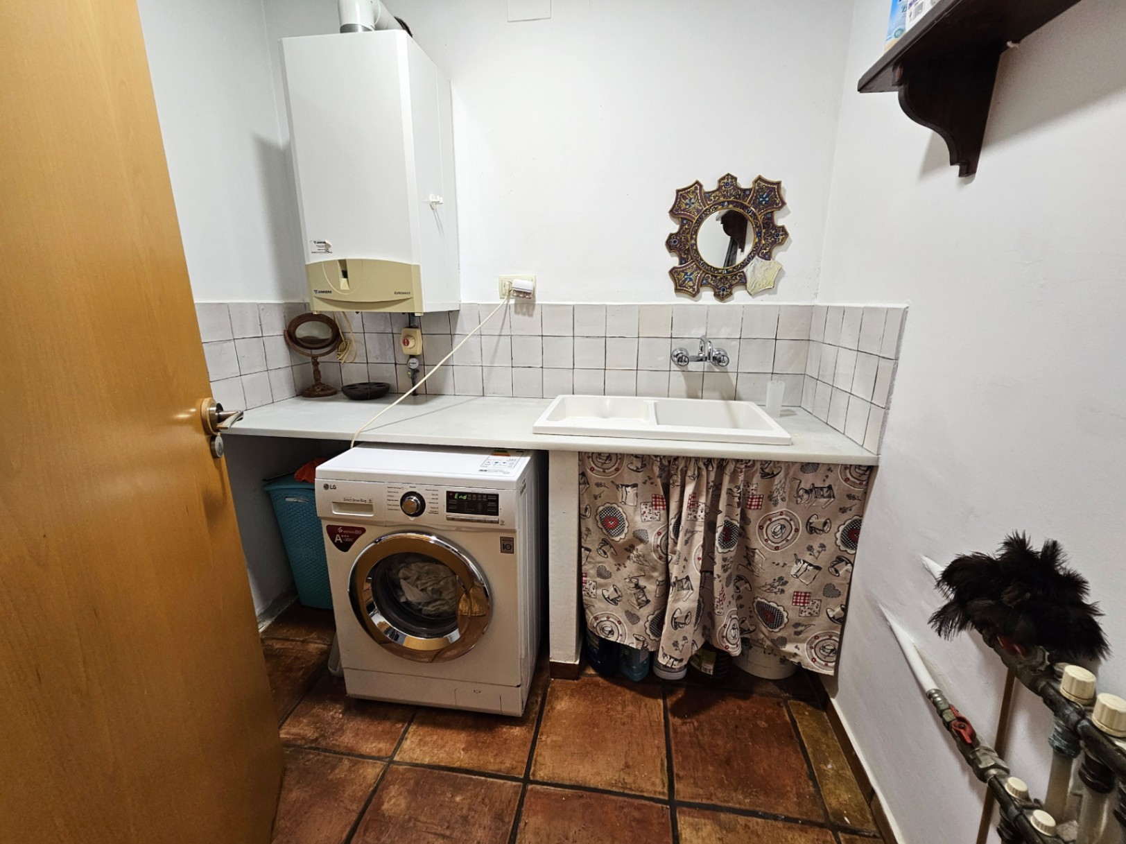 Cuarto de lavado funcional con lavadora LG, encimera blanca con fregadero doble y suelos de barro manual. Incluye caldera mural y revestimiento cerámico blanco.