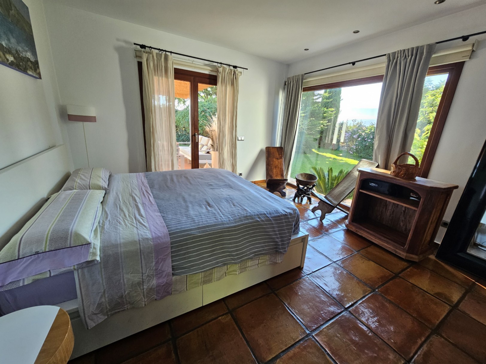 Dormitorio luminoso con suelos de barro hechos a mano, amplios ventanales con vistas al jardín y acceso directo a la zona exterior.