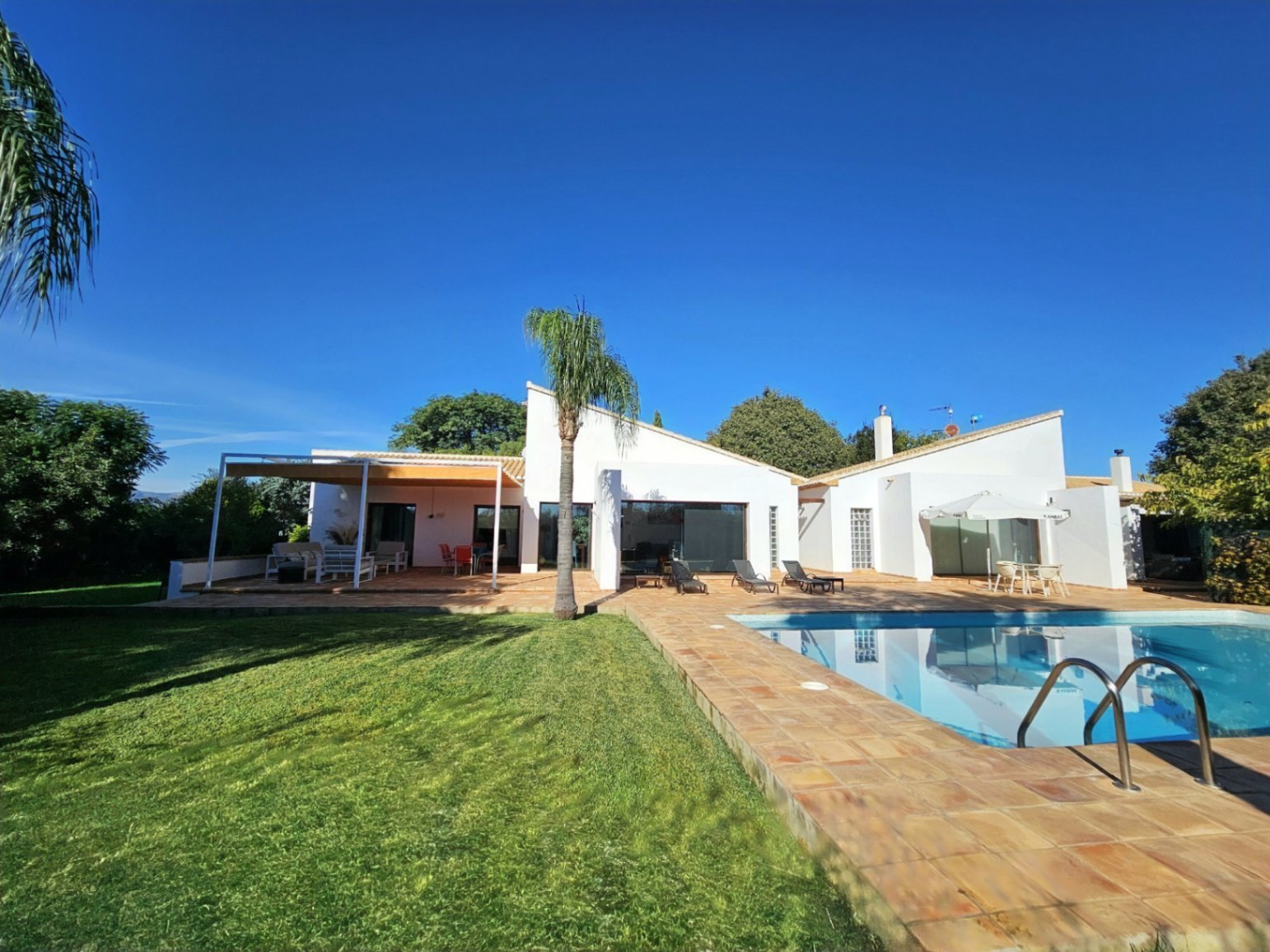 Exterior de la villa con piscina, rodeada de suelos de barro hechos a mano, amplias zonas de césped y una terraza cubierta que conecta con el interior.