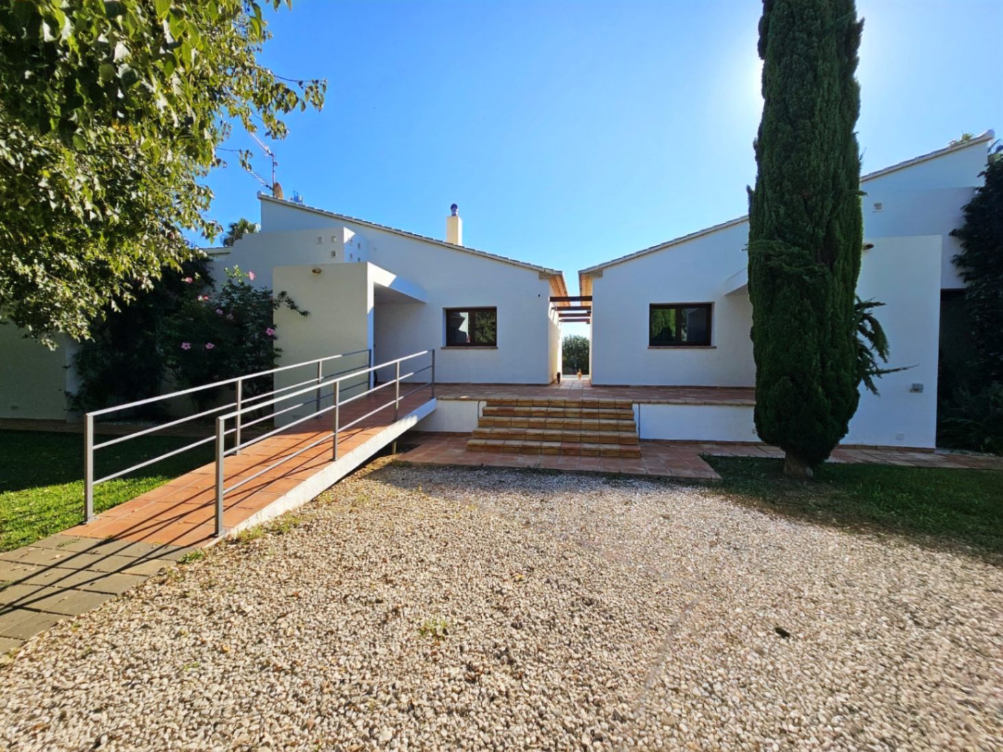 Fachada exterior de las dos viviendas independientes con acceso mediante rampa metálica y escaleras de barro, rodeadas de jardines y zona de grava.