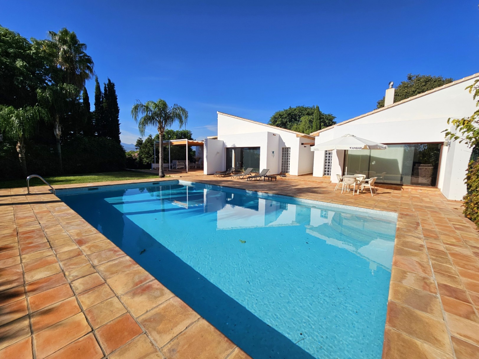 Piscina rectangular rodeada de una terraza de barro artesanal, con vistas a la fachada blanca de la villa y jardines mediterráneos bajo un cielo despejado.