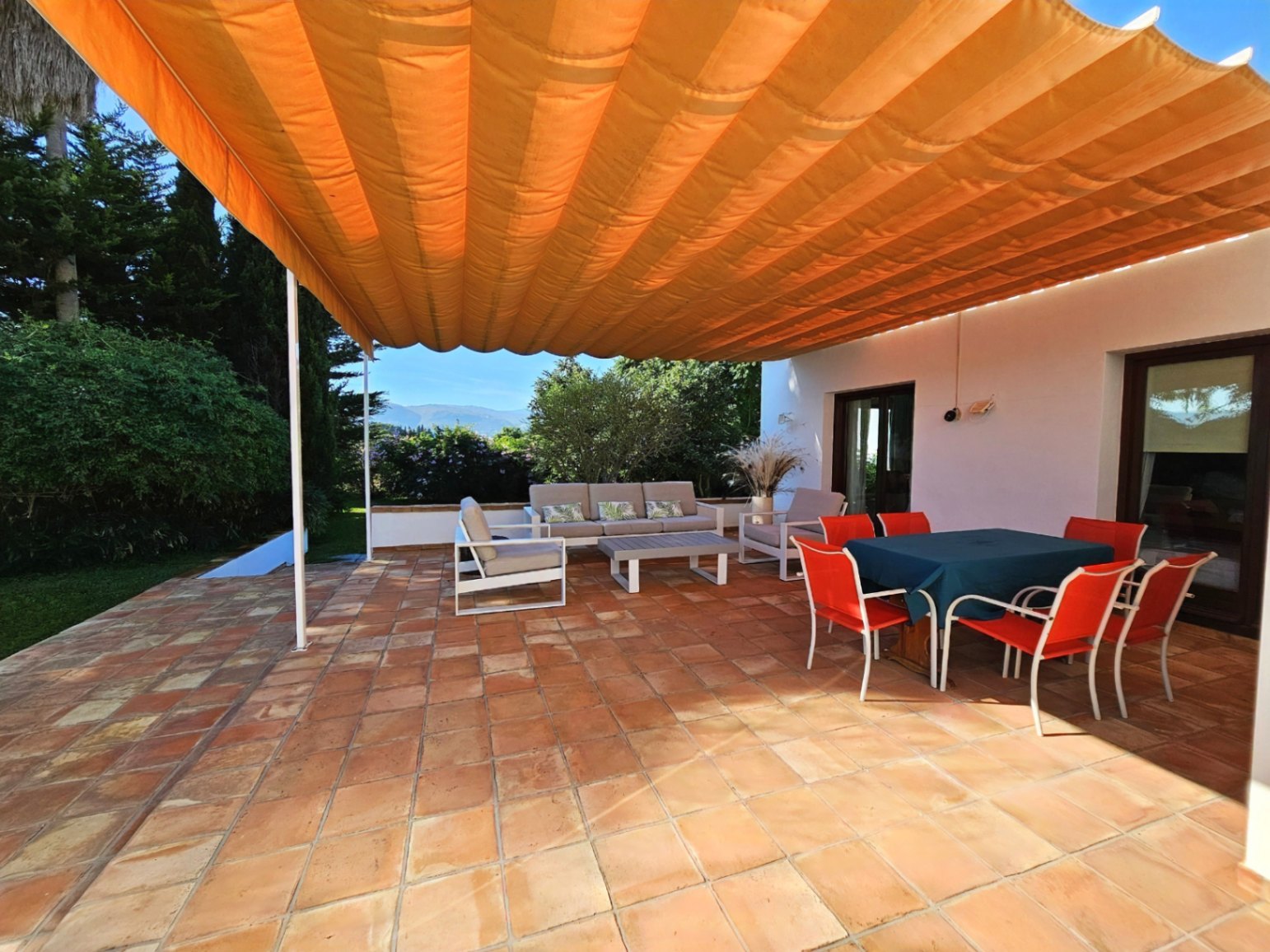 Terraza exterior con suelos de barro manual, gran toldo naranja retráctil y zonas de comedor y descanso rodeadas de vegetación mediterránea.