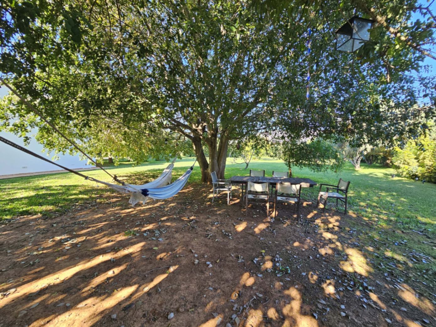 Zona de descanso sombreada bajo un gran árbol con hamaca y mesa de comedor exterior, rodeada de un extenso jardín con césped.