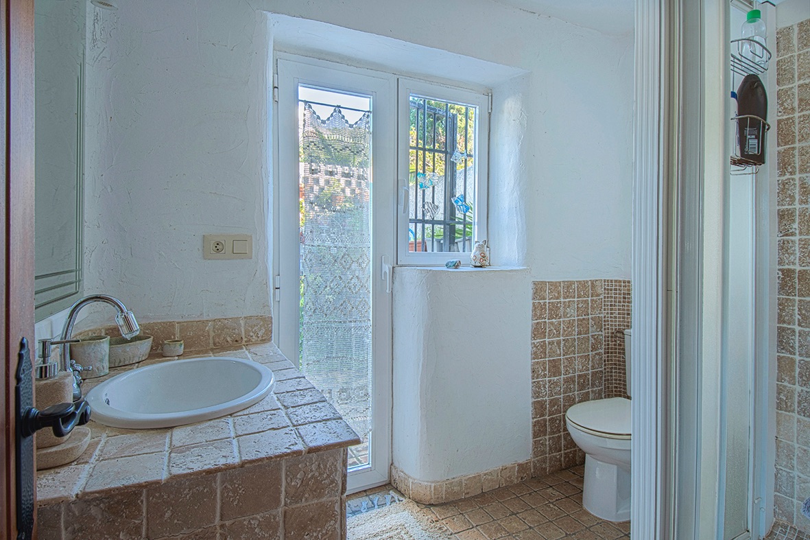 Baño de estilo rústico con encimera de azulejos artesanales, lavabo encastrado y paredes de yeso texturizado. Incluye ventana de climalit con reja de seguridad.