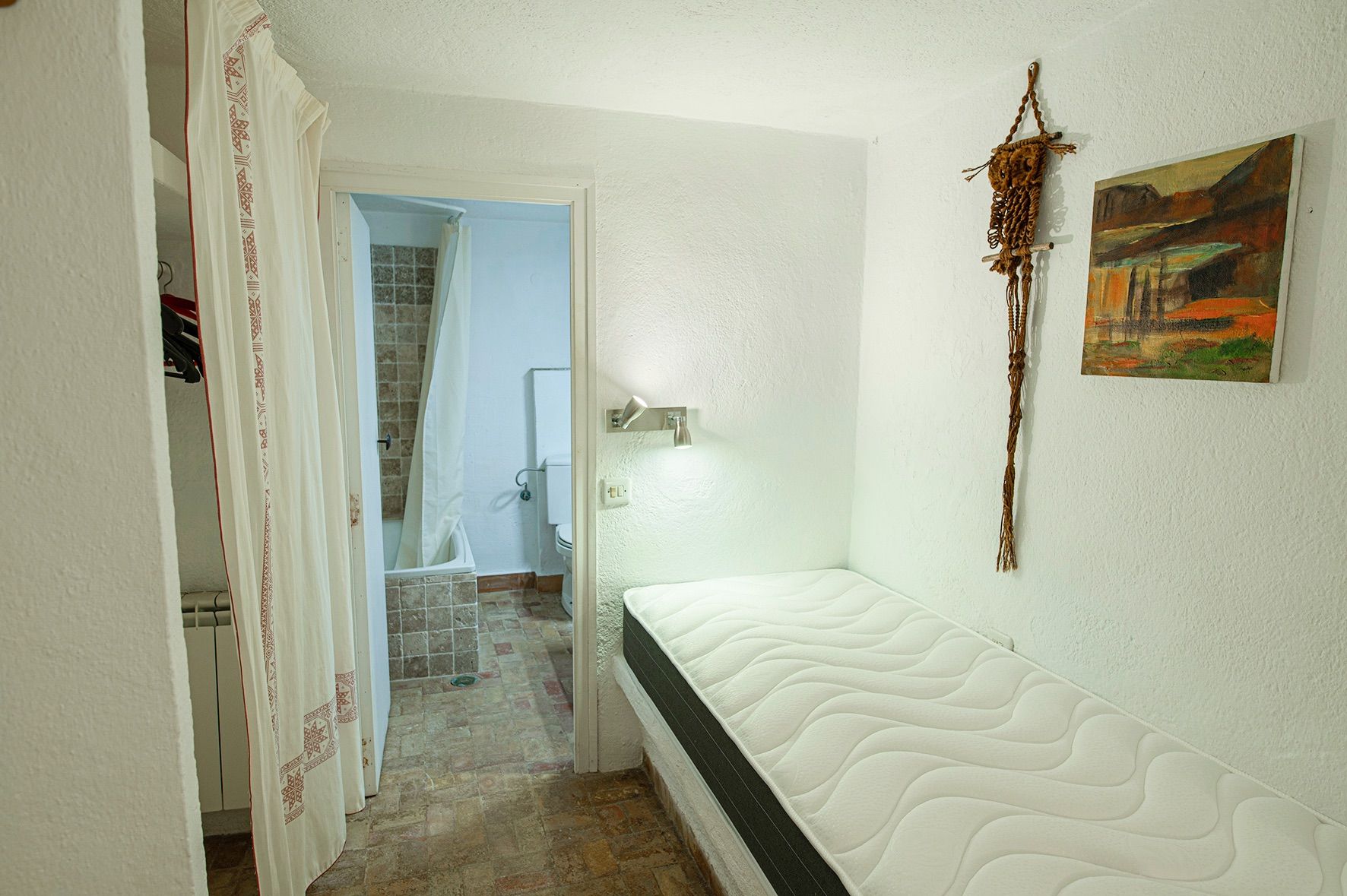 Dormitorio con cama individual y baño en suite, suelos de barro cocido y paredes blancas de estilo rústico tradicional.