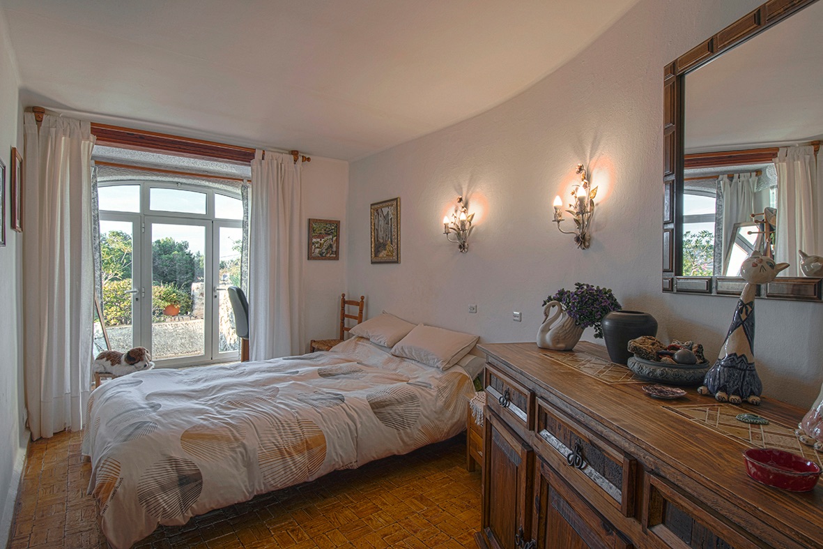 Dormitorio de estilo tradicional con suelos de barro, mobiliario de madera rústica y acceso directo al exterior mediante una puerta acristalada de arco.