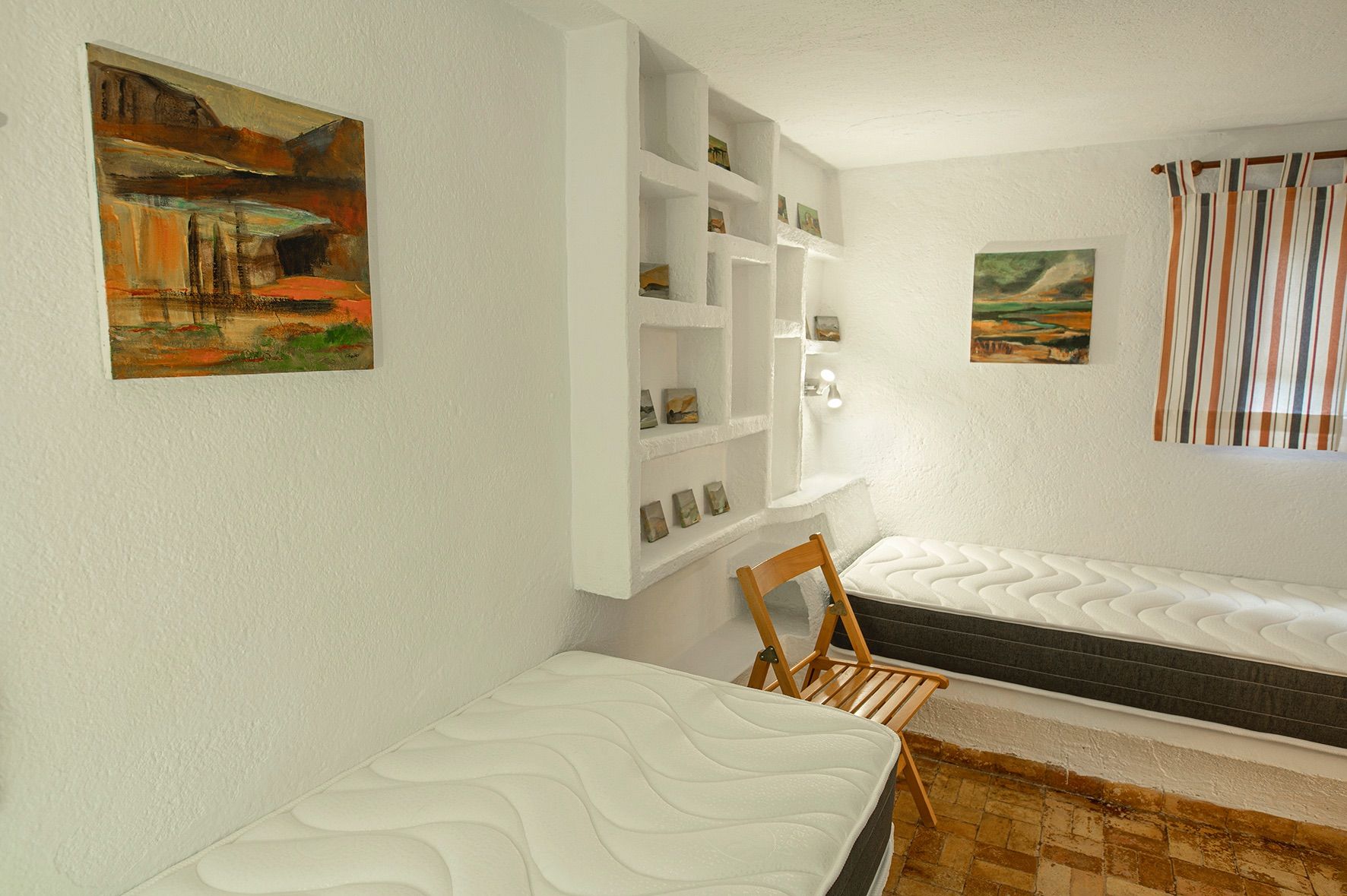 Dormitorio rústico con dos camas individuales, estantería de obra integrada en la pared blanca y suelo de baldosas de barro cocido.