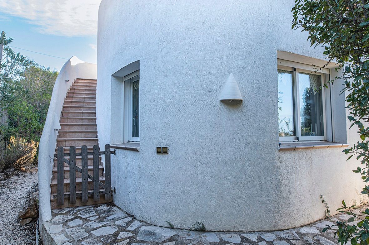 Fachada curva de estilo mediterráneo con paredes de estuco blanco, escalera exterior con peldaños cerámicos y acceso mediante una pequeña puerta de madera.