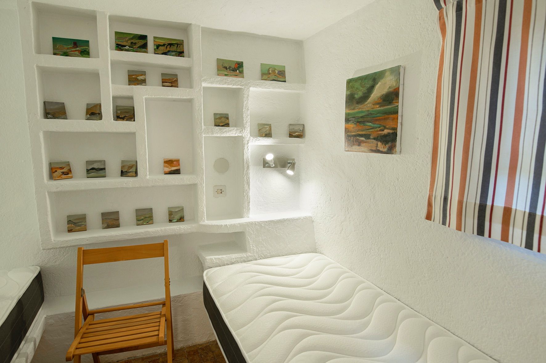 Habitación con paredes blancas de mampostería y nichos integrados que exhiben pequeñas pinturas, complementada con una cama individual y una silla de madera.