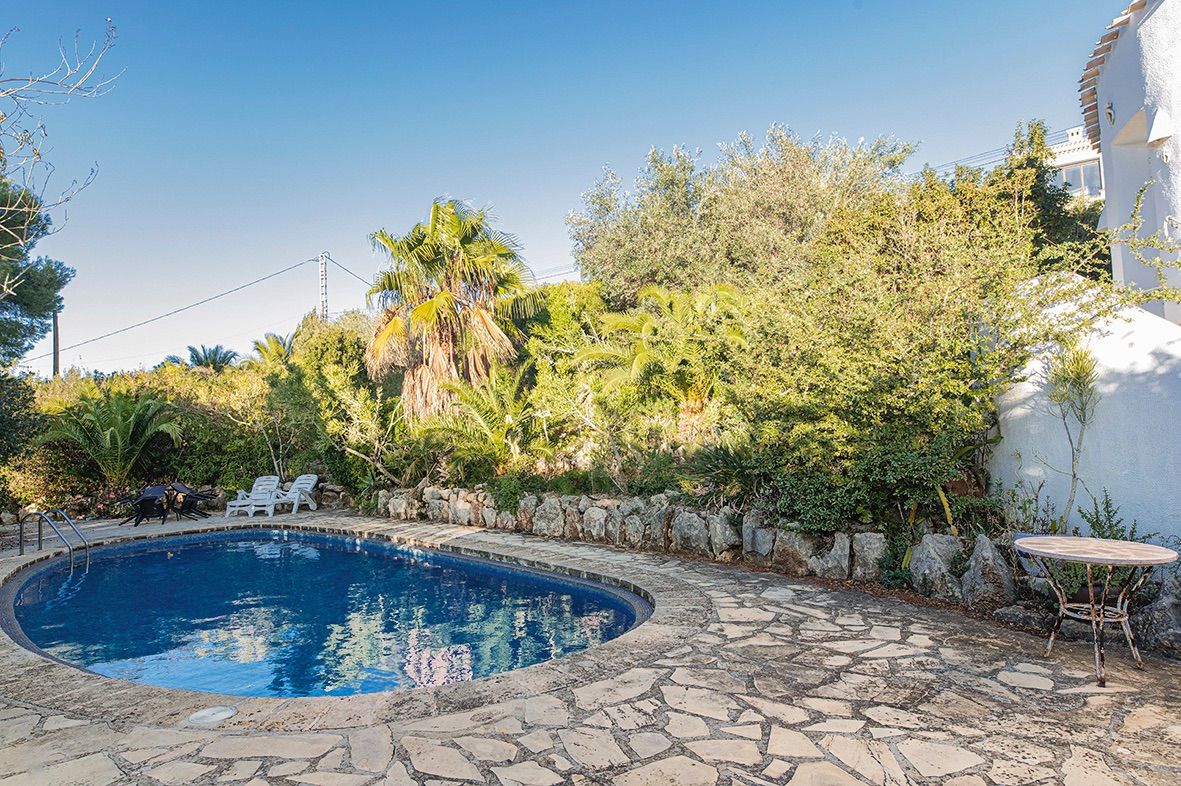 Piscina de forma riñonada rodeada por una terraza de piedra natural y un jardín mediterráneo maduro con palmeras y muros de piedra rústica.