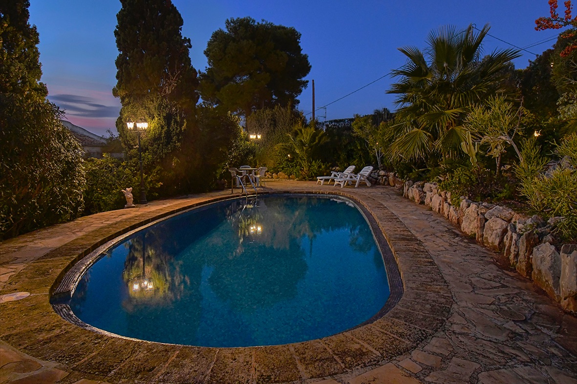 Piscina exterior iluminada rodeada de pavimento de piedra natural y un jardín mediterráneo con palmeras y cipreses bajo el cielo del atardecer.