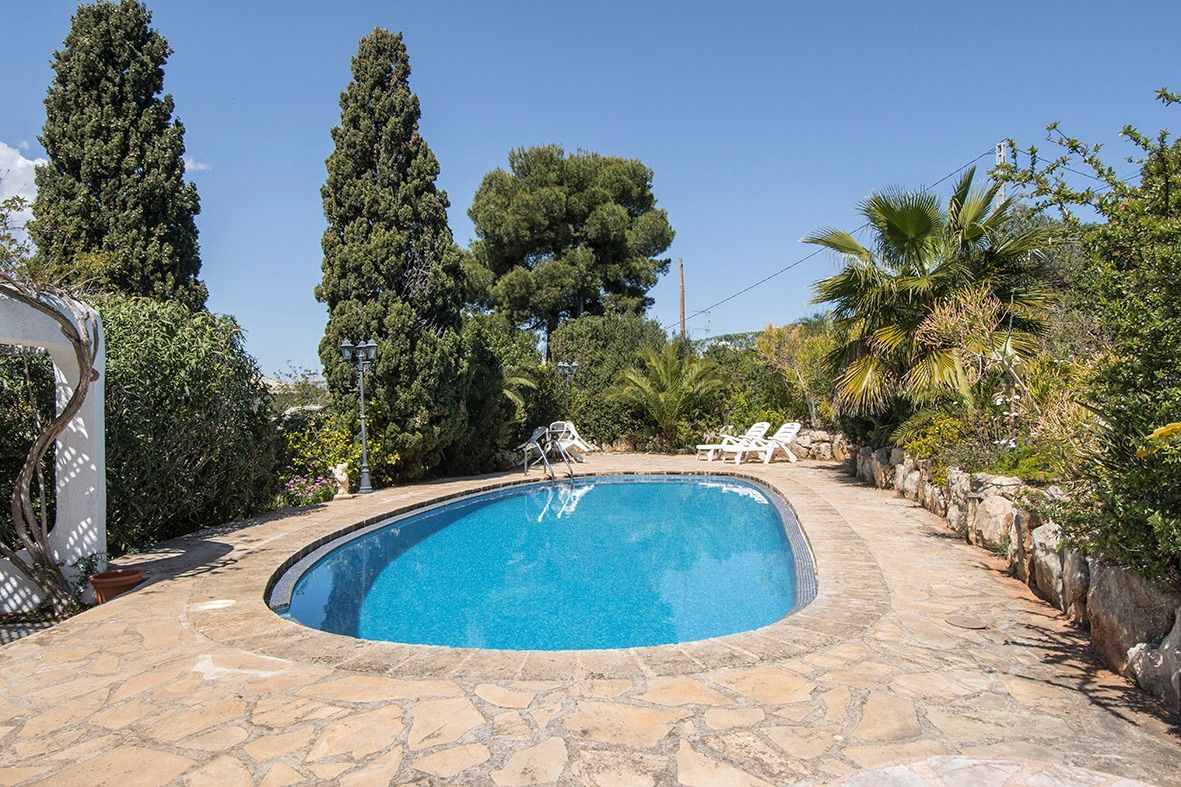 Piscina ovalada rodeada de una terraza de piedra natural y un jardín mediterráneo maduro con cipreses y palmeras.