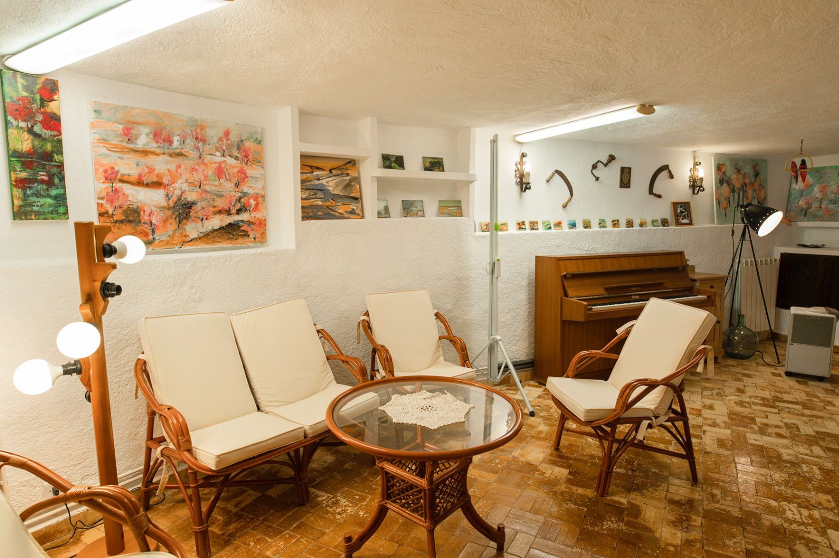 Sala de estar rústica con muebles de mimbre, piano de pared y suelos de terracota. Las paredes blancas exhiben diversas obras de arte y nichos integrados.