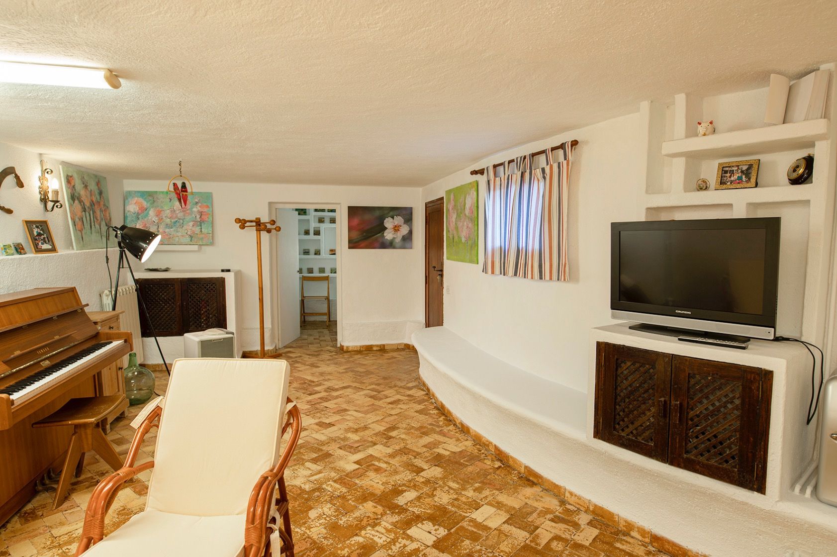 Sala de estar rústica con suelos de barro cocido, un piano de pared de madera y mobiliario integrado en mampostería blanca con puertas de celosía.