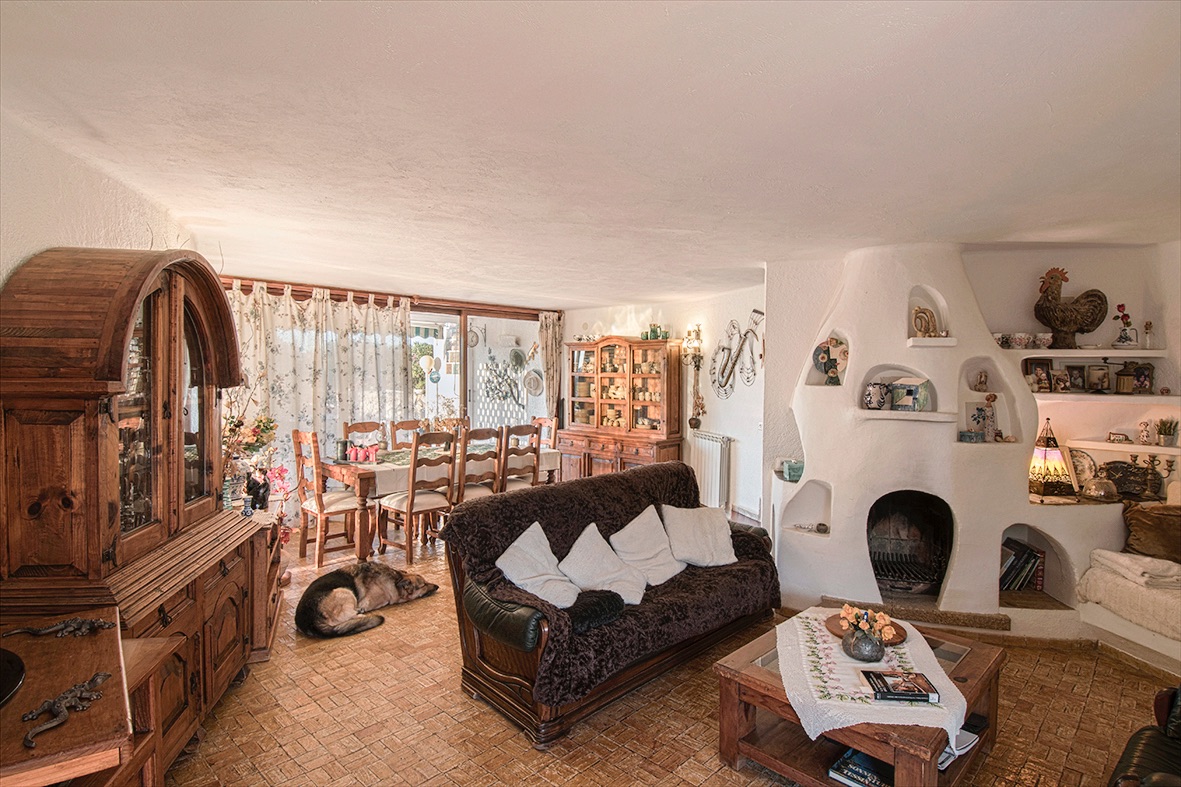 Salón comedor rústico con chimenea de obra blanca esculpida, muebles de madera maciza, suelos de gres y acceso a la terraza a través de amplios ventanales.