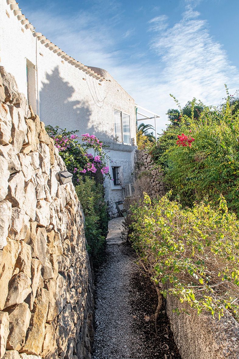 Sendero exterior flanqueado por muros de piedra natural y vegetación mediterránea, que conduce a la fachada blanca de estilo tradicional con detalles de buganvilla.