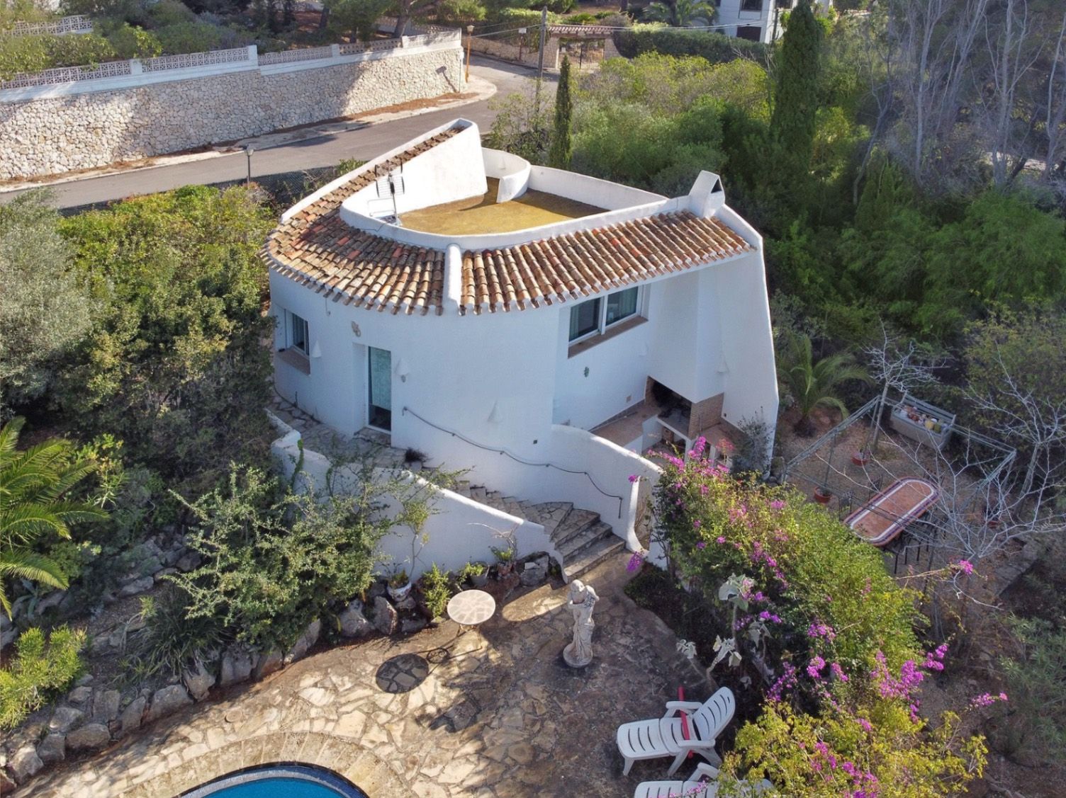 Vista aérea de la villa secundaria con arquitectura curva, zona de barbacoa integrada, escaleras de piedra y patio pavimentado junto a la vegetación mediterránea.