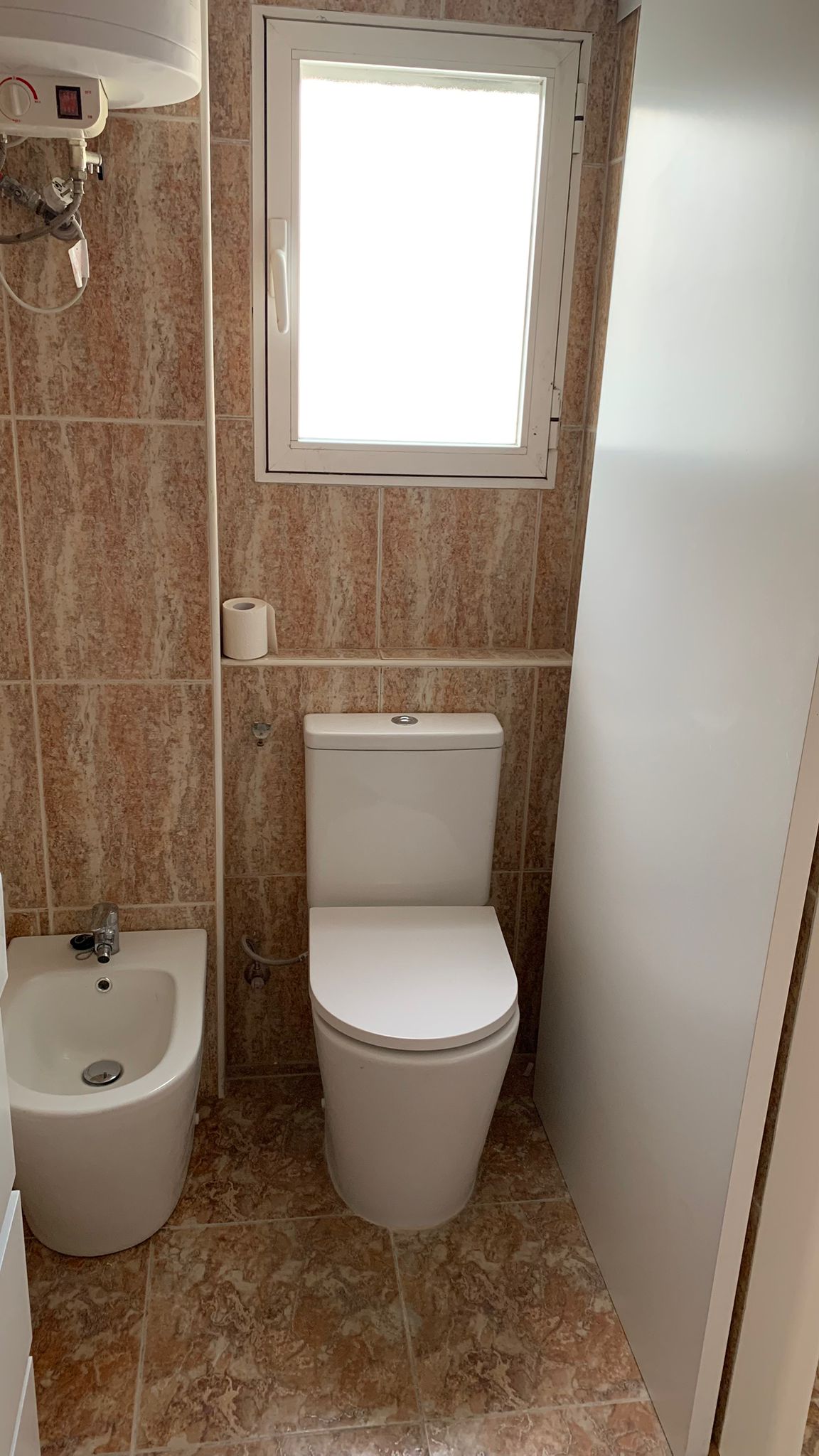 Cuarto de baño con inodoro y bidé de porcelana blanca, revestido con azulejos de tonos tierra y una ventana que aporta luz natural y ventilación.