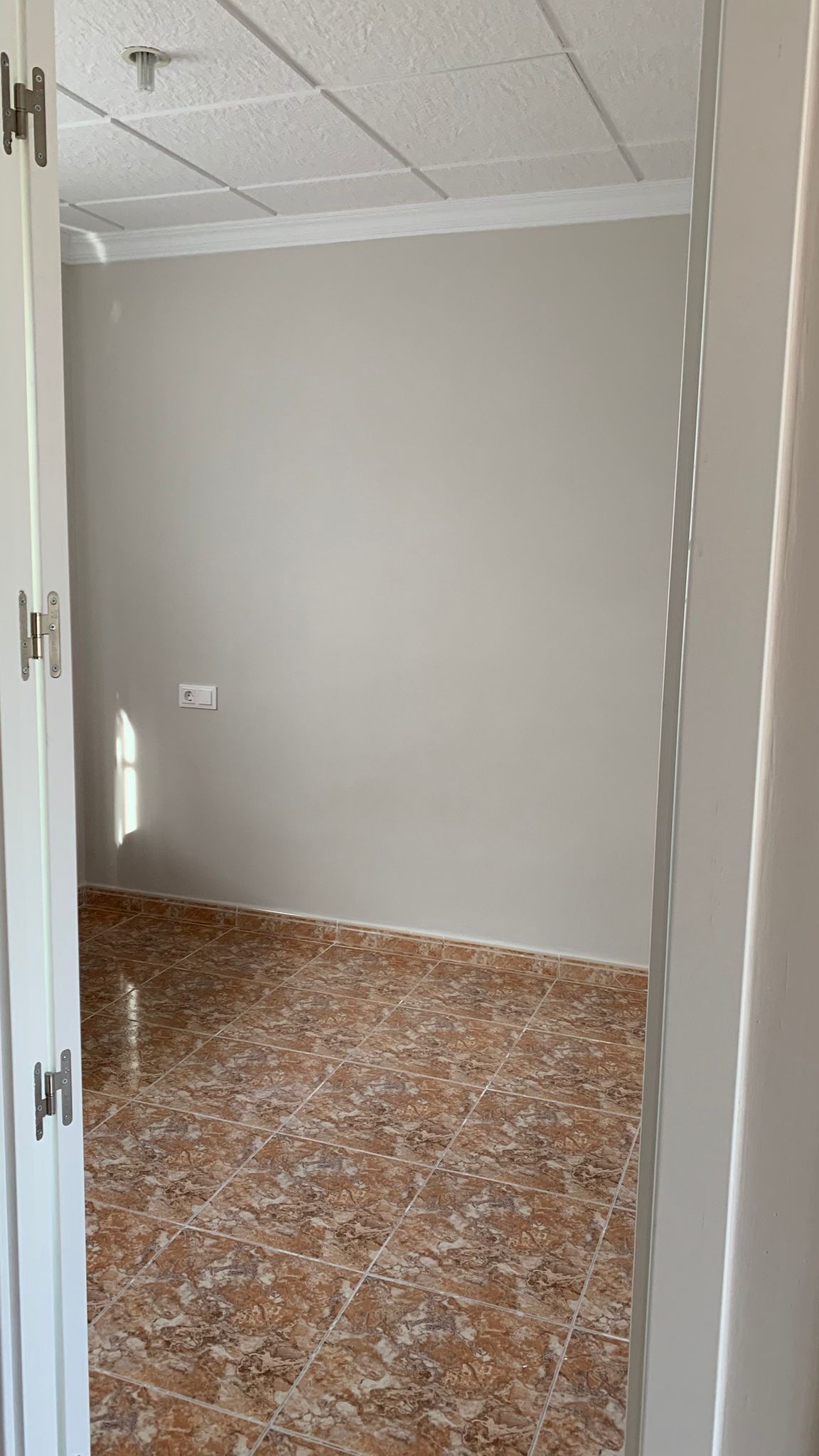 Habitación vacía con suelos de gres cerámico en tonos marrones, paredes en gris suave y techo técnico blanco. La estancia cuenta con iluminación natural lateral.