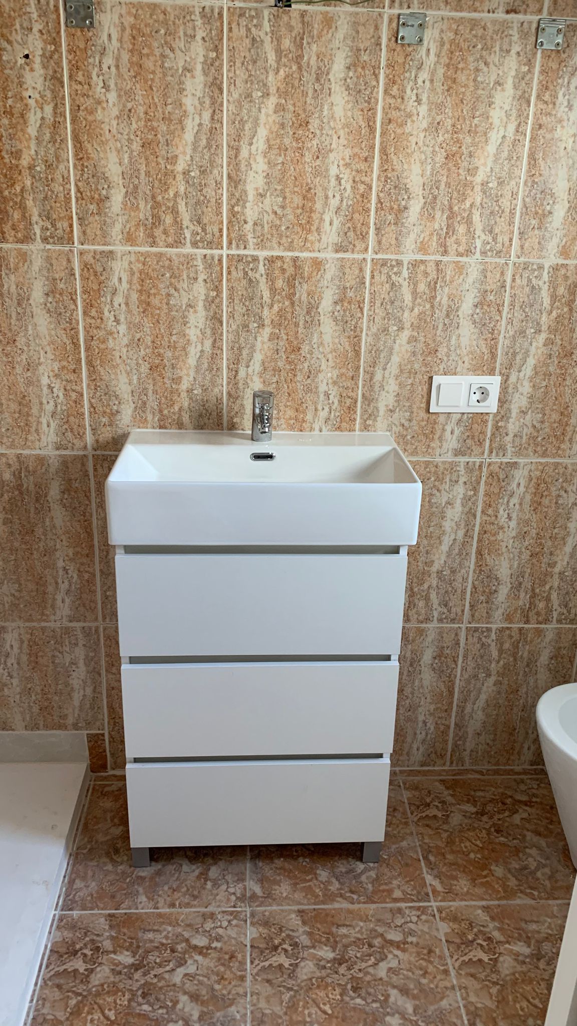 Mueble de baño moderno en blanco con tres cajones y lavabo integrado, rodeado de azulejos cerámicos en tonos tierra con efecto mármol.