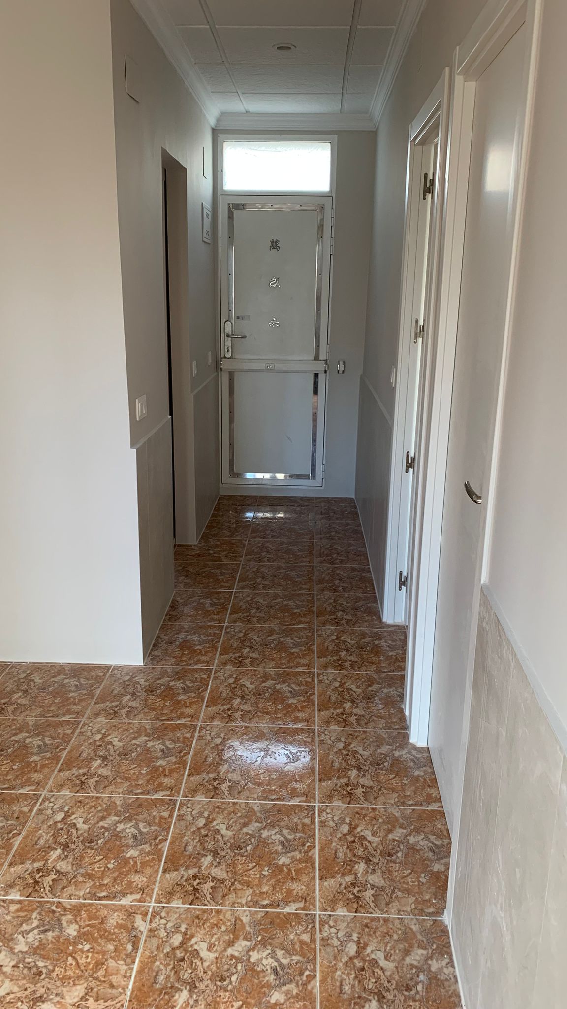 Pasillo de entrada con suelos cerámicos marmoleados en tonos marrones y paredes blancas. Al fondo se aprecia una puerta metálica de seguridad con un tragaluz superior.