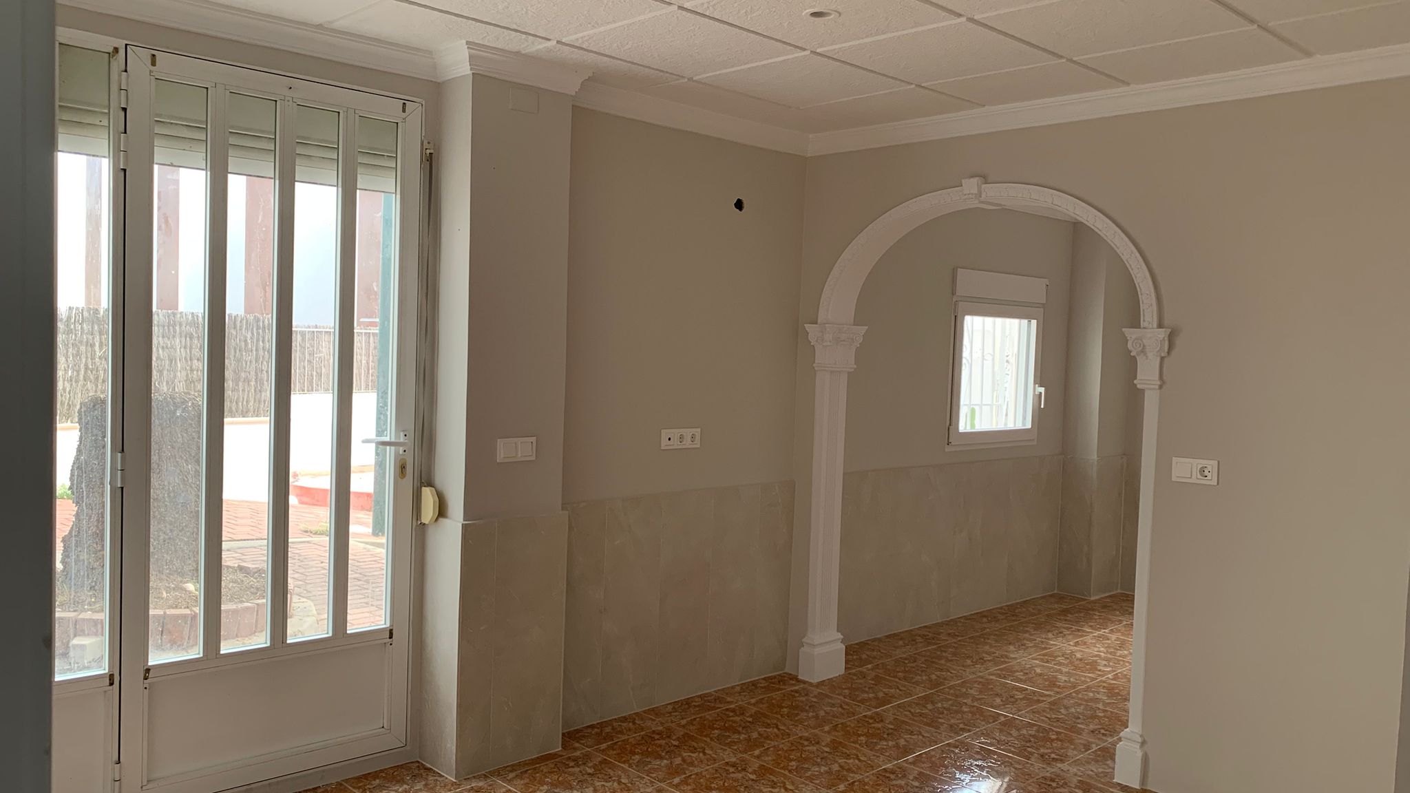 Salón con suelos cerámicos, paredes de tono neutro y un arco decorativo blanco. Incluye acceso directo al exterior mediante una puerta acristalada con rejas de seguridad.
