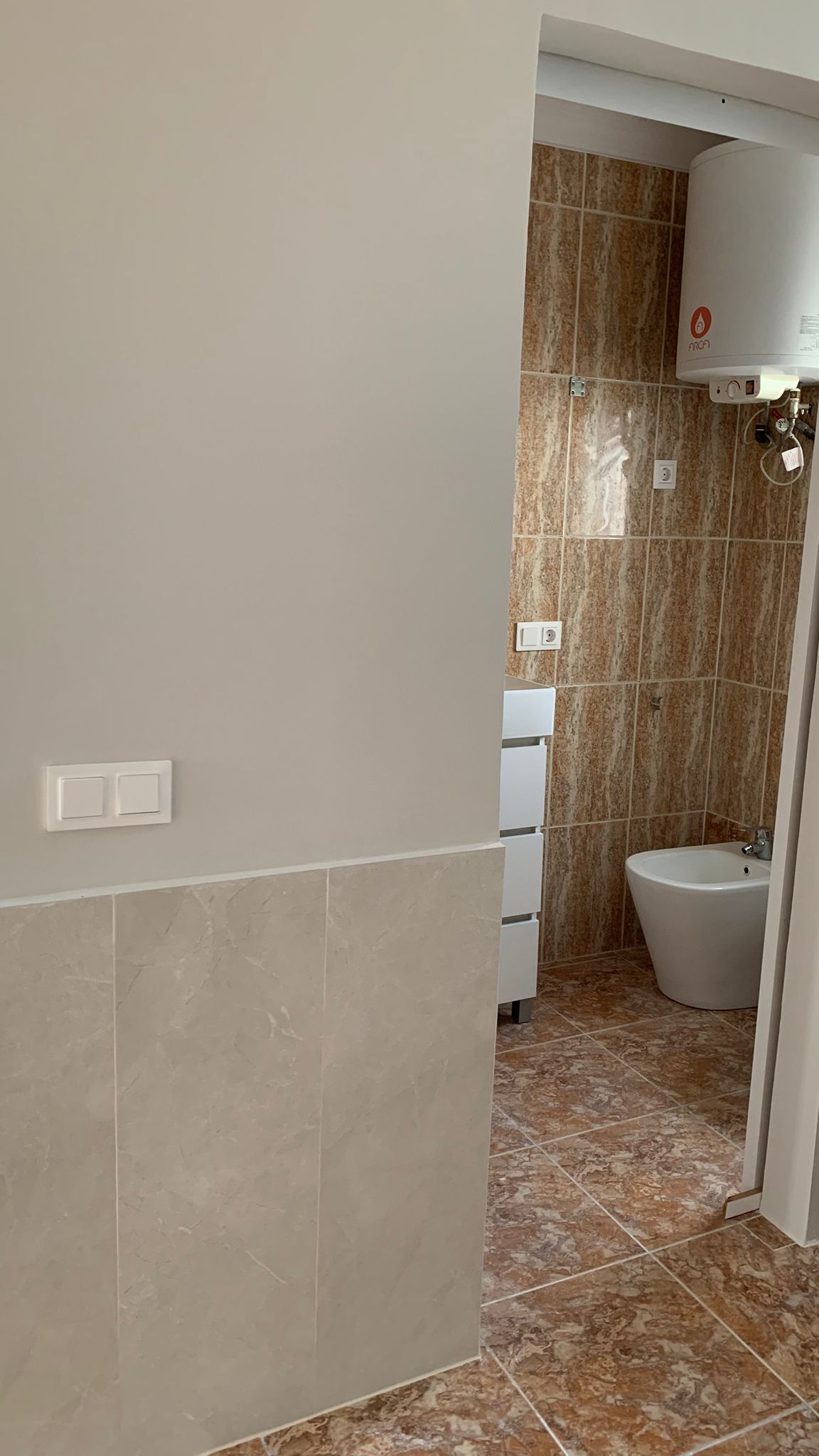 Vista del baño con bidé, mueble auxiliar blanco y termo eléctrico, revestido con azulejos de tonos marrones y veteado marmóreo.