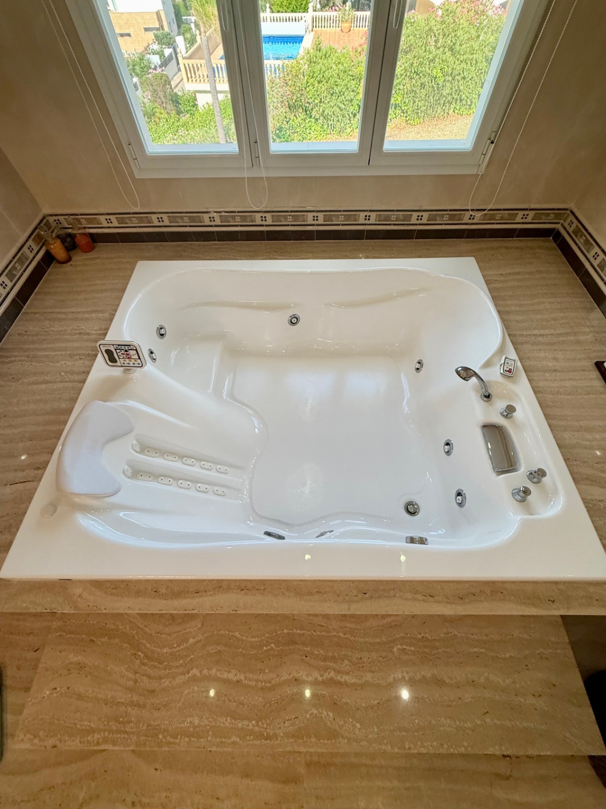 Bañera de hidromasaje integrada en mármol travertino, equipada con reposacabezas y controles digitales, situada frente a un ventanal con vistas al jardín y la piscina.