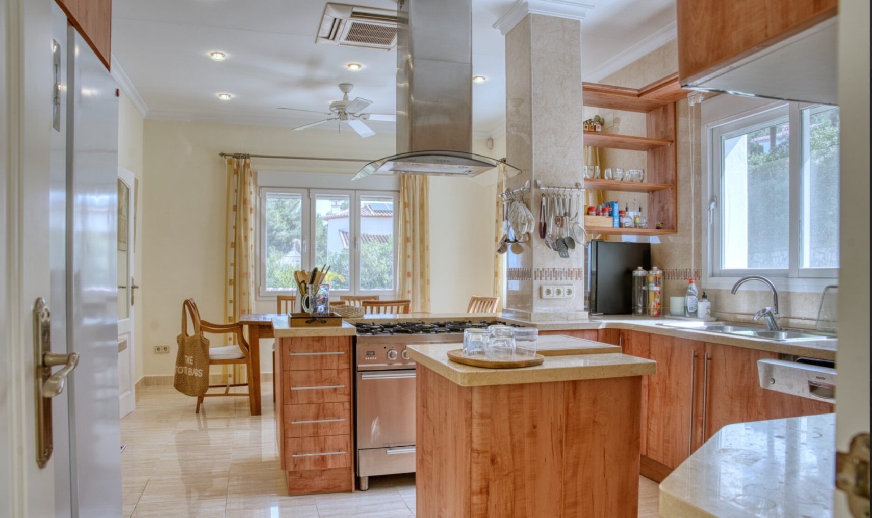 Cocina equipada con mobiliario de madera, encimeras de piedra clara y una isla central con fogones de gas y campana extractora de acero inoxidable.
