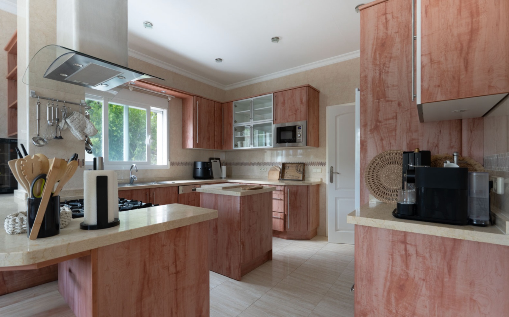 Cocina independiente con mobiliario de madera, encimeras de piedra natural e isla central. Dispone de electrodomésticos de acero inoxidable y luz natural a través de su amplio ventanal.