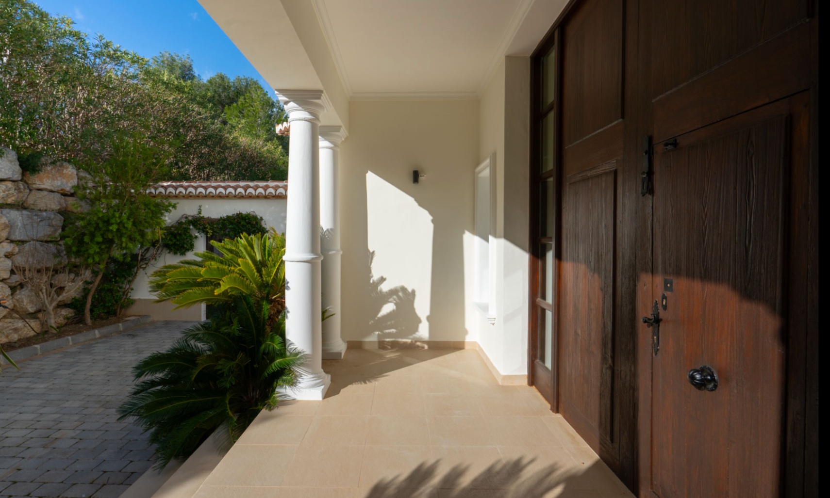 Entrada principal con puerta de madera maciza, columnas blancas clásicas y porche sombreado rodeado de vegetación mediterránea y muros de piedra natural.