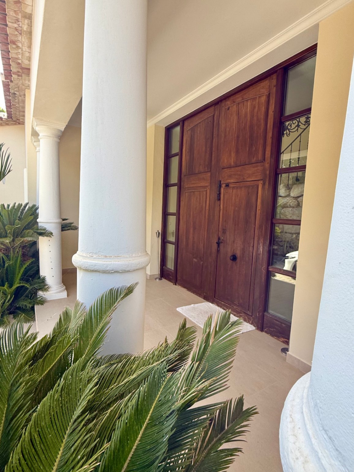 Entrada principal con puerta doble de madera maciza, flanqueada por ventanales laterales y columnas blancas de estilo toscano en un porche rodeado de vegetación mediterránea.