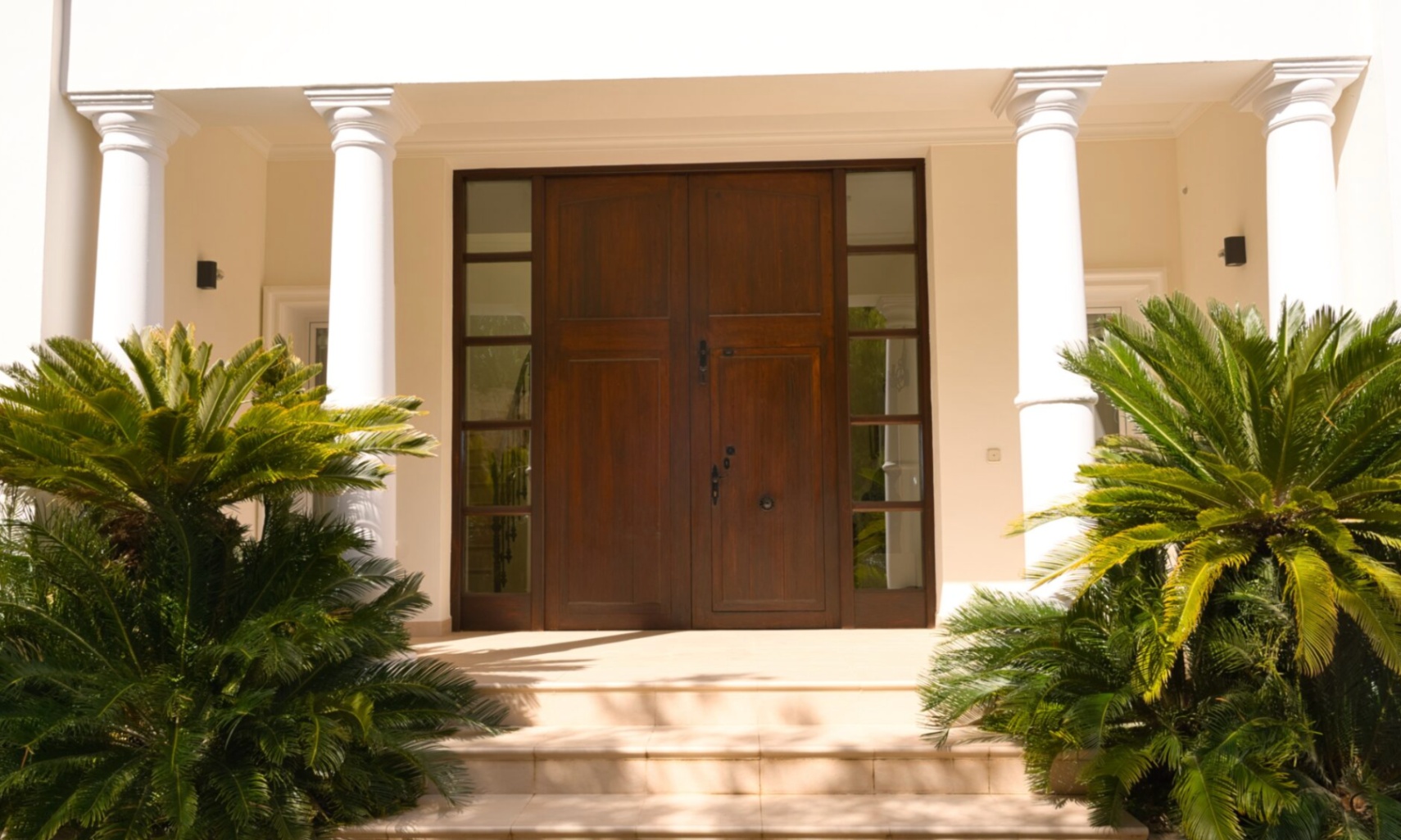 Entrada principal con puerta doble de madera oscura y ventanales laterales, enmarcada por columnas blancas clásicas y plantas de cica.