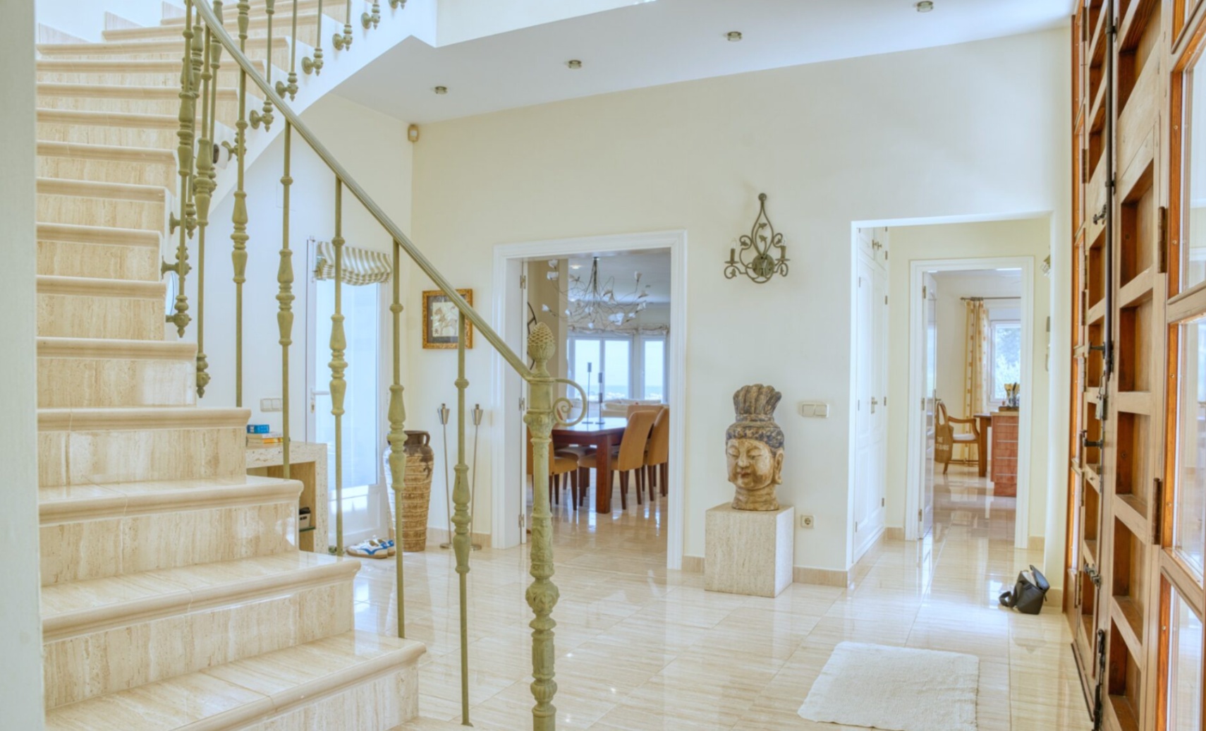 Hall de entrada con escalera de mármol travertino y barandilla de hierro forjado. Suelos de piedra pulida conectan con el comedor y ofrecen vistas al mar al fondo.