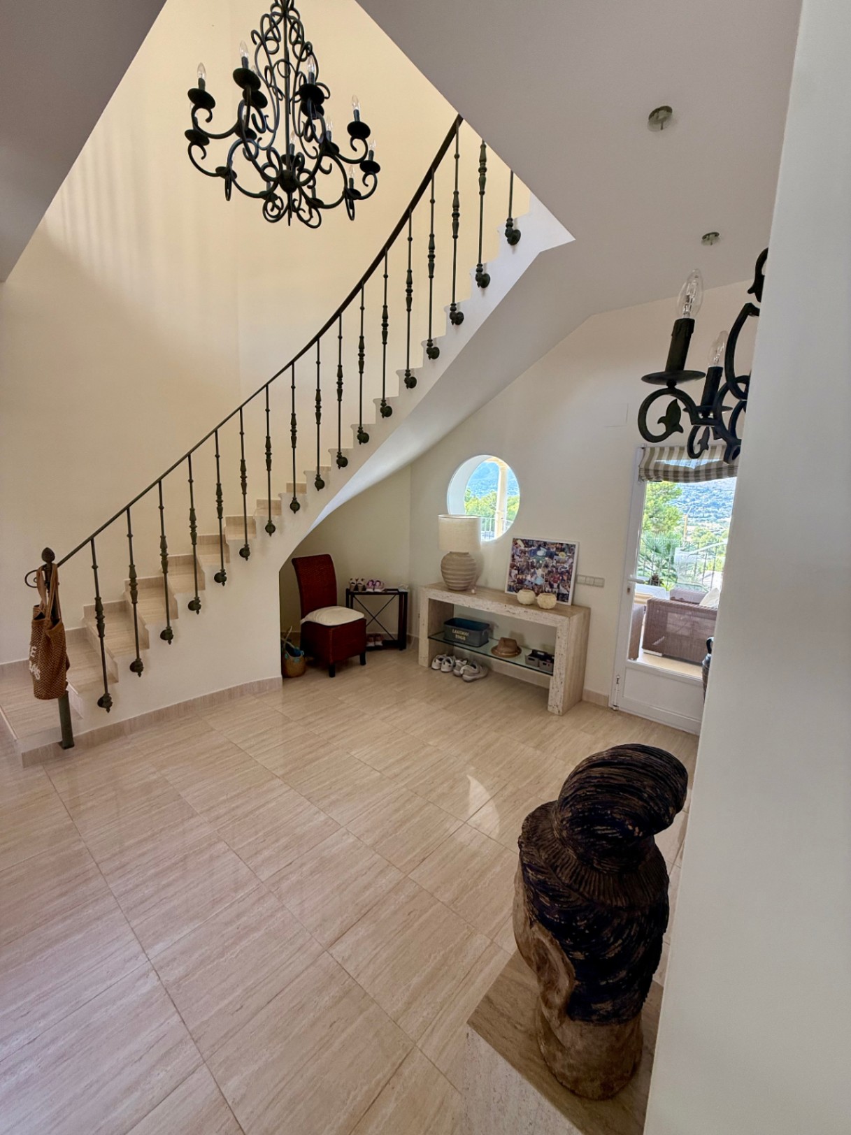 Hall de entrada con escalera de mármol y barandilla de hierro forjado. Destaca la lámpara de araña clásica, el suelo de piedra pulida y una ventana circular decorativa.