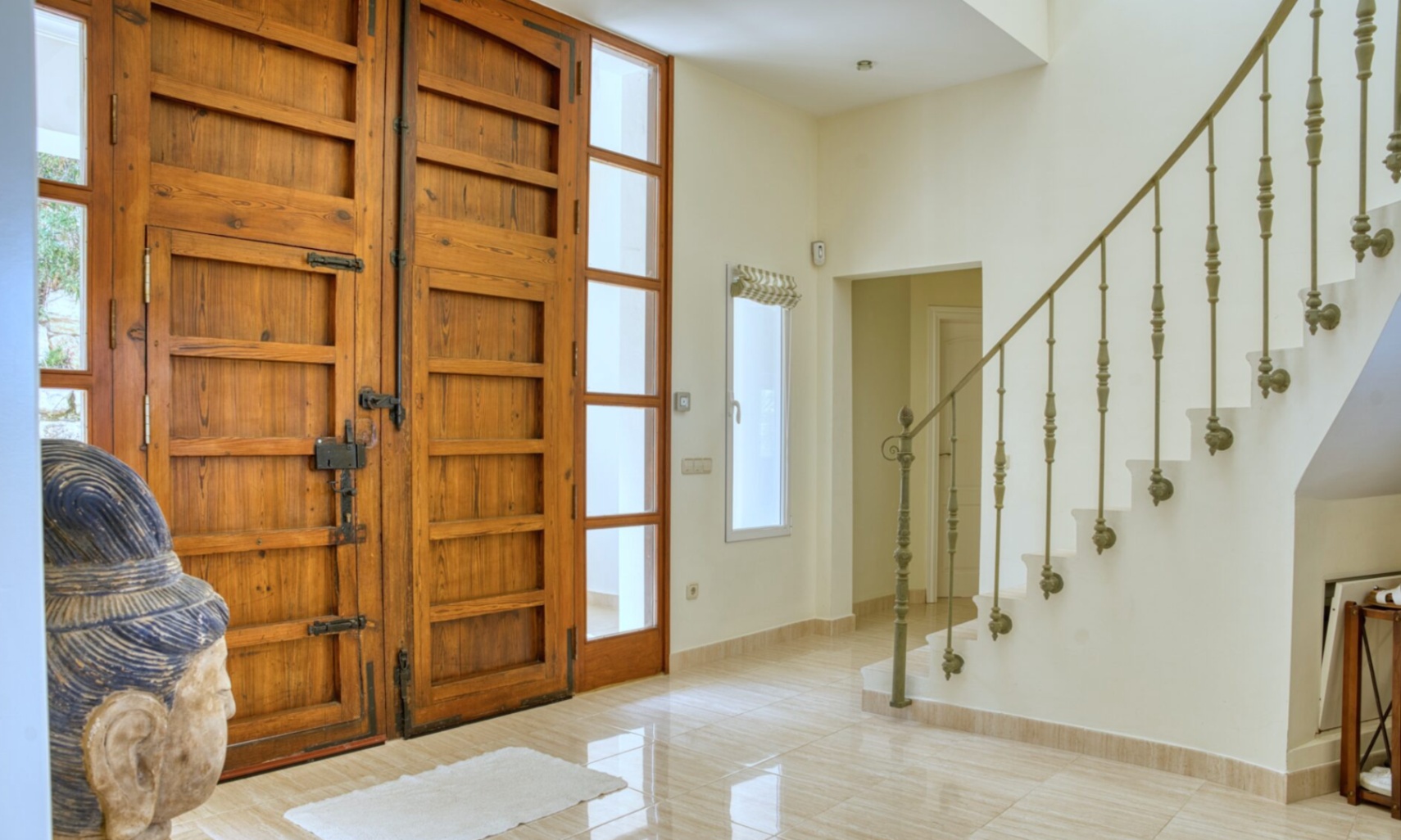 Hall de entrada con gran puerta doble de madera rústica, herrajes de hierro y escalera de mármol con barandilla ornamental sobre suelos cerámicos pulidos.