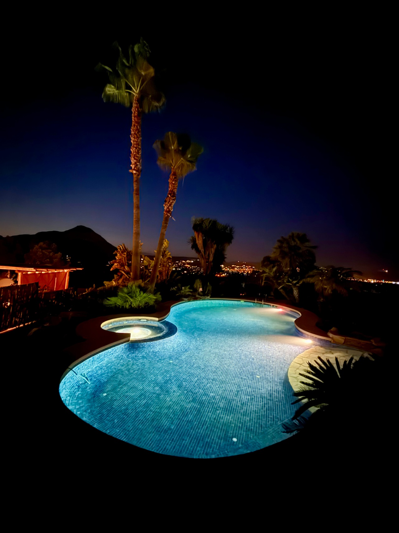 Piscina de diseño curvo con iluminación nocturna y jacuzzi integrado, rodeada de palmeras con vistas a las luces de la ciudad y la silueta del Montgó.