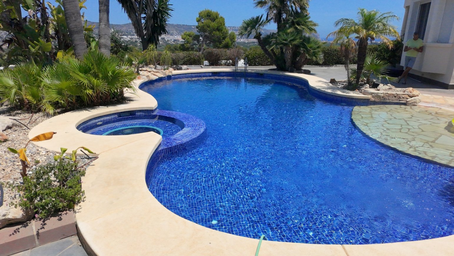 Piscina de forma orgánica con revestimiento de mosaico azul, jacuzzi circular integrado y terraza de piedra rodeada de palmeras y vegetación mediterránea.