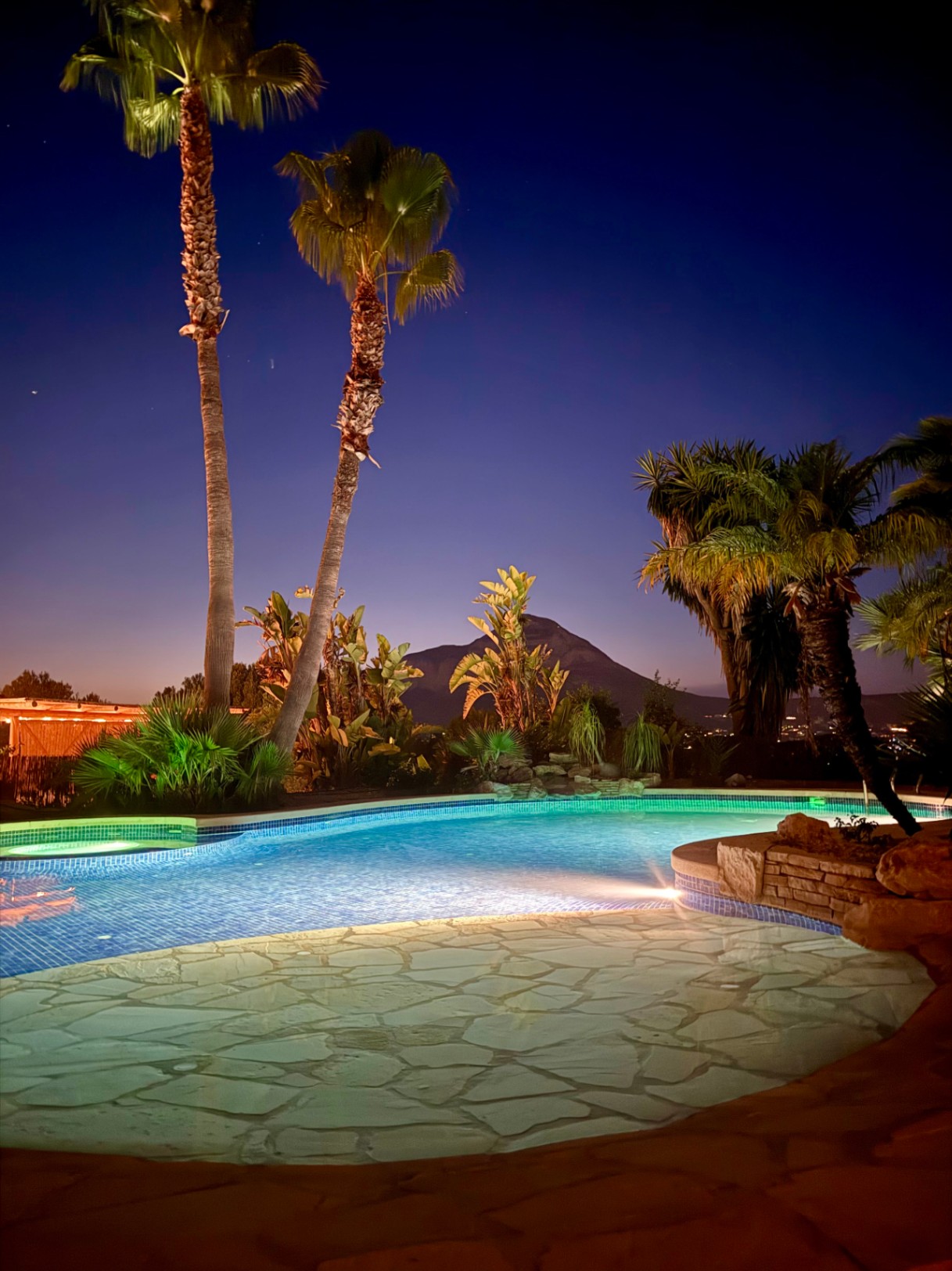 Piscina iluminada de noche con revestimiento de piedra natural y vistas a la silueta del Montgó, rodeada de palmeras y vegetación tropical.