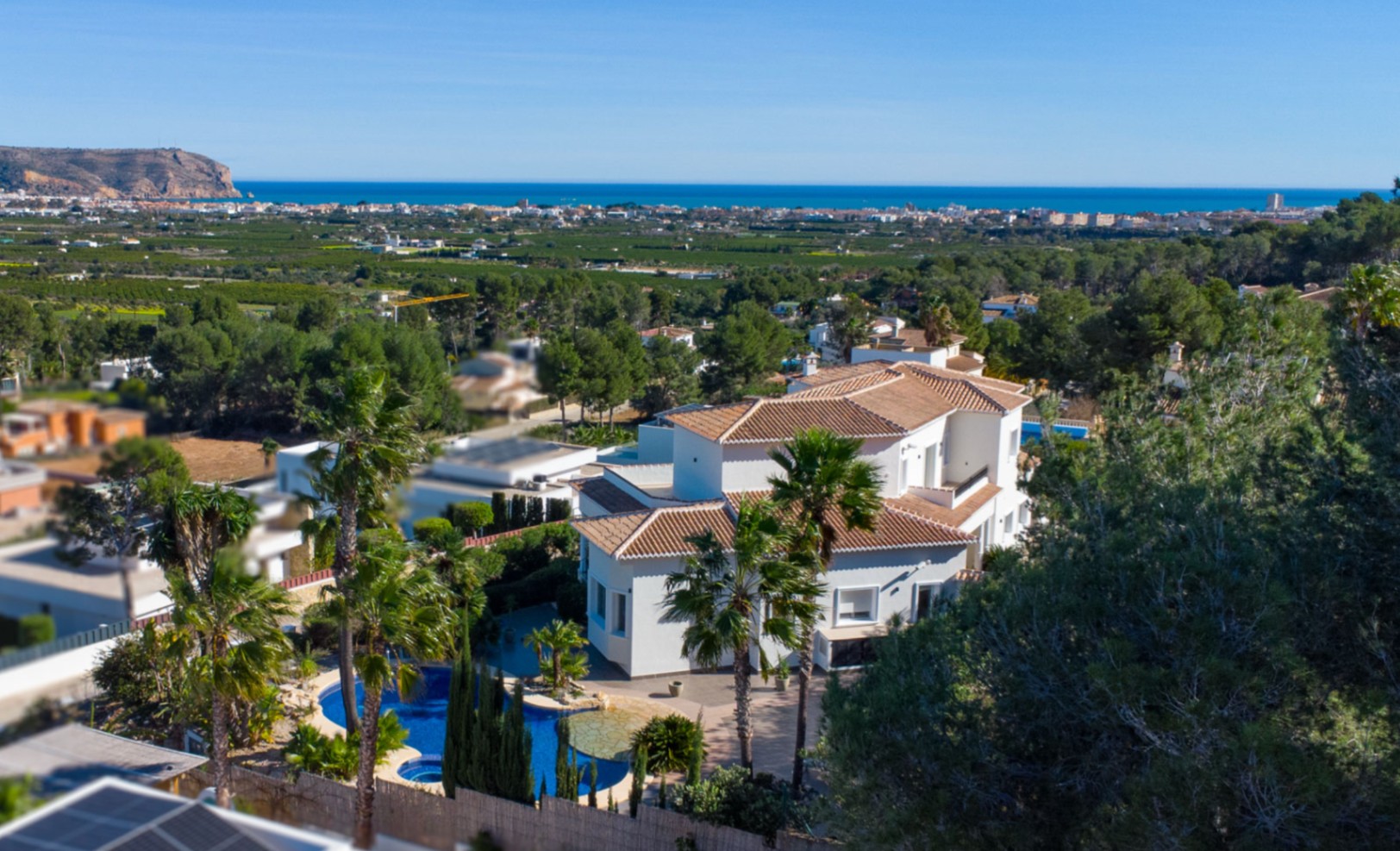 Vista aérea de la villa blanca con tejados de teja, destacando la piscina orgánica y el extenso jardín con palmeras frente al mar Mediterráneo.
