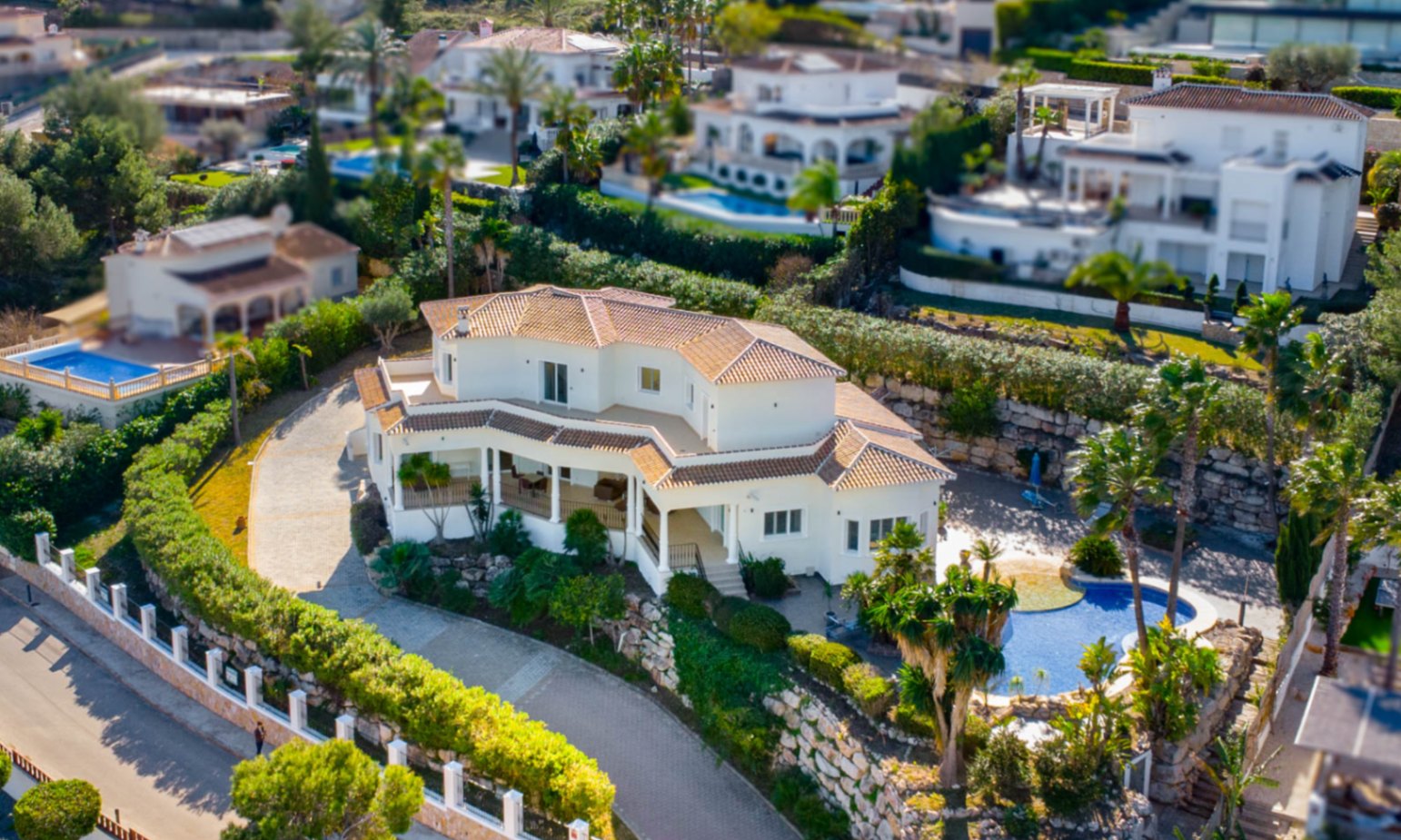 Vista aérea de la villa con fachada blanca y tejado de teja cerámica, destacando su piscina de diseño orgánico y el acceso pavimentado rodeado de vegetación mediterránea.